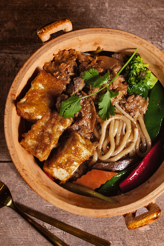 Tontopf mit Nudeln, Fleisch, Tofu und Gemüse; braune, warme Farben.