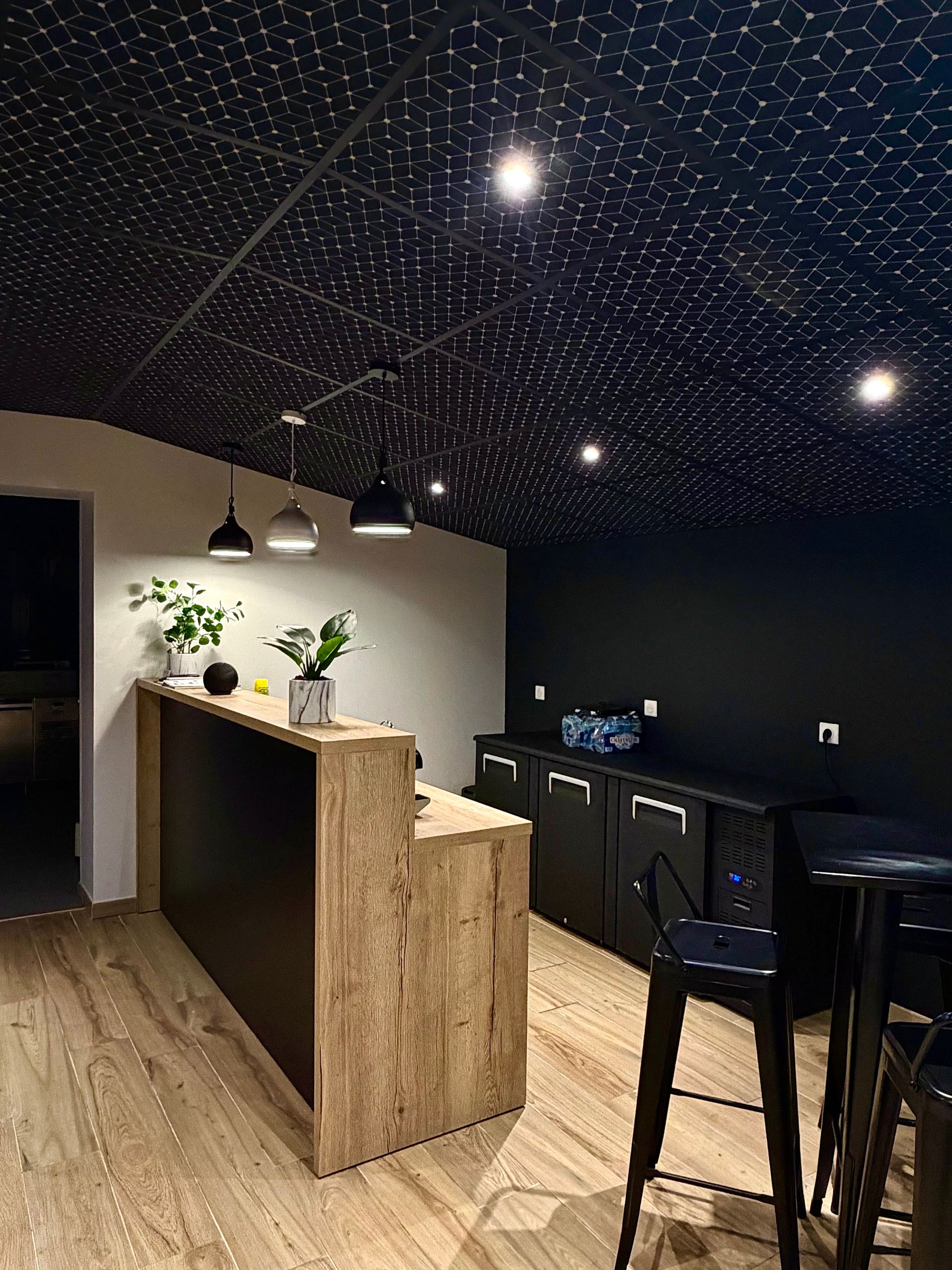Un espace d'accueil moderne avec un comptoir en bois clair, des murs noirs et un plafond étoilé orné de suspensions lumineuses.