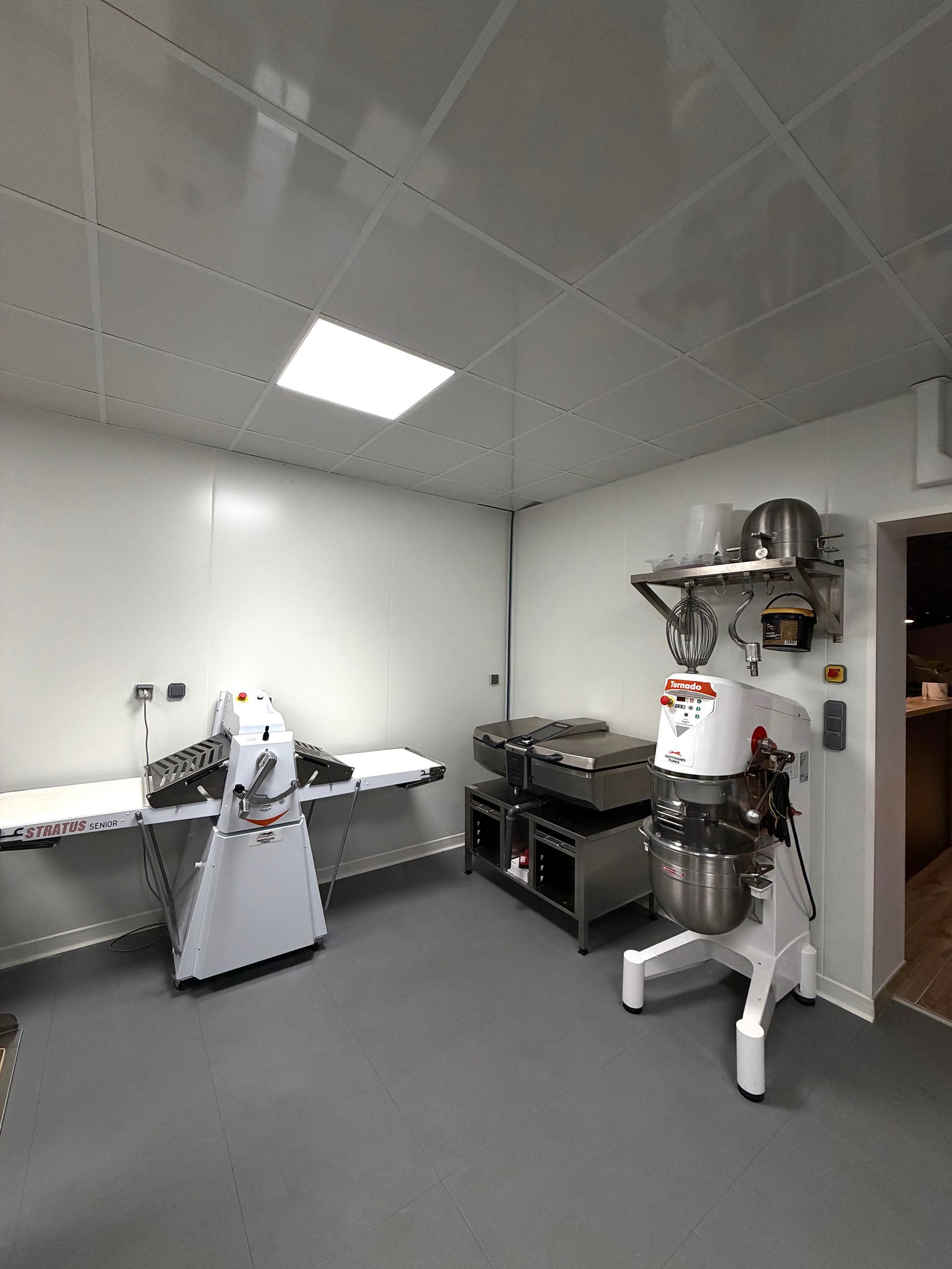 Une cuisine professionnelle comprenant une laminoir à pâte, une table de travail en acier inoxydable et un grand batteur sur socle.