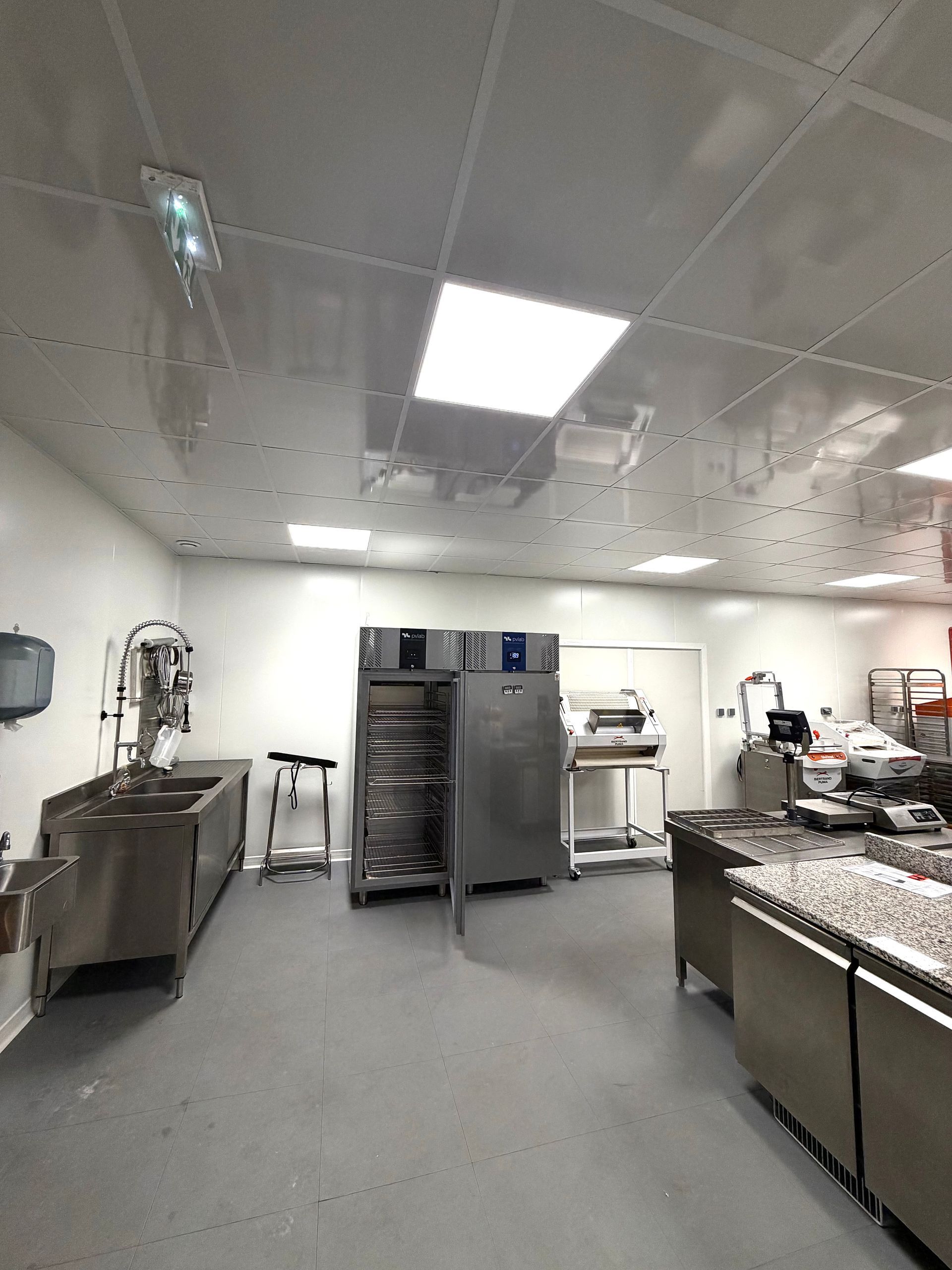 Une cuisine professionnelle équipée de matériel en acier inoxydable, de tables de préparation, d'une étagère métallique et d'un éclairage lumineux au plafond.