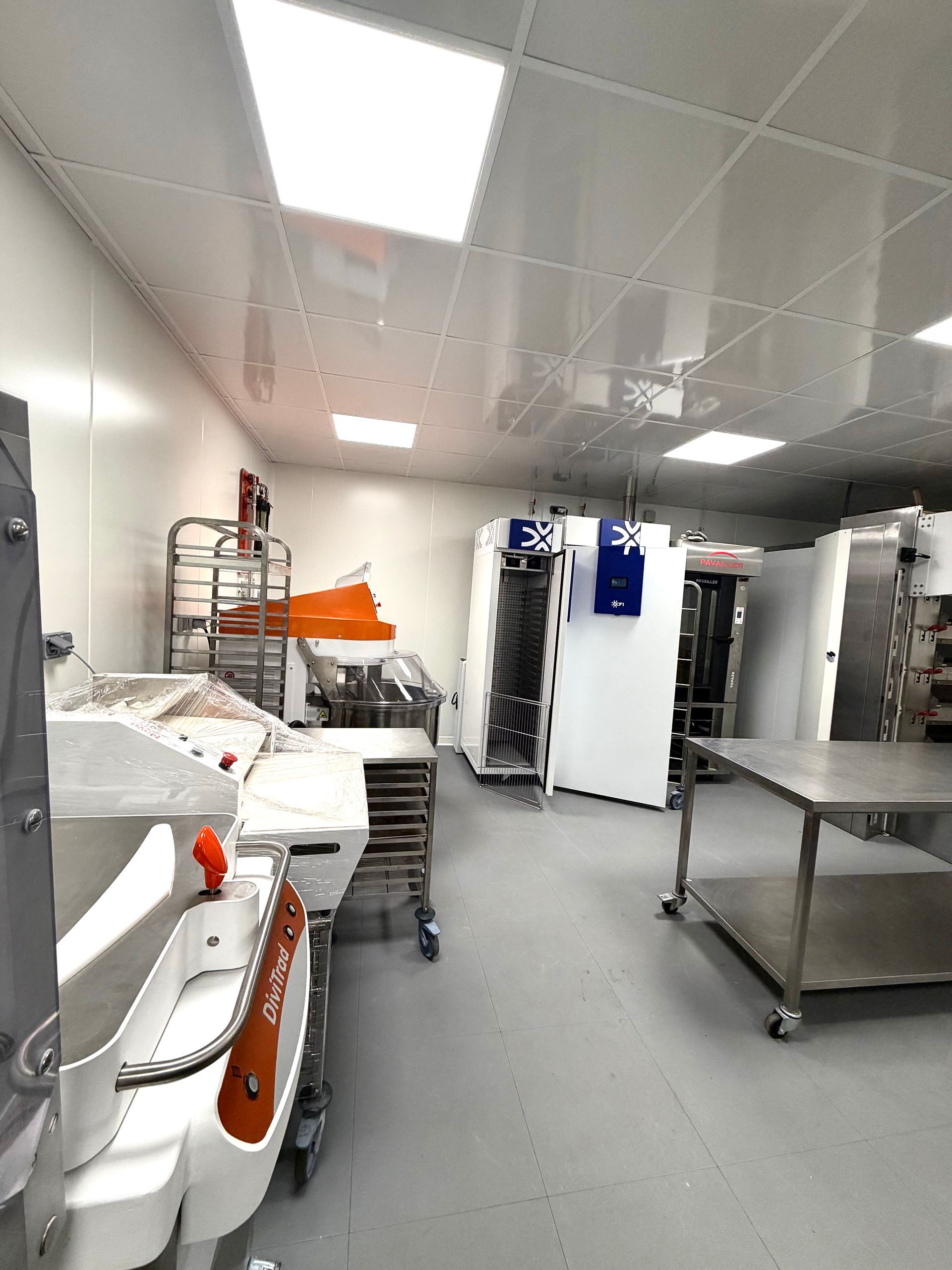 Une cuisine industrielle bien éclairée, équipée de grandes machines à pâtisserie blanches, de tables en acier inoxydable et d'étagères métalliques.