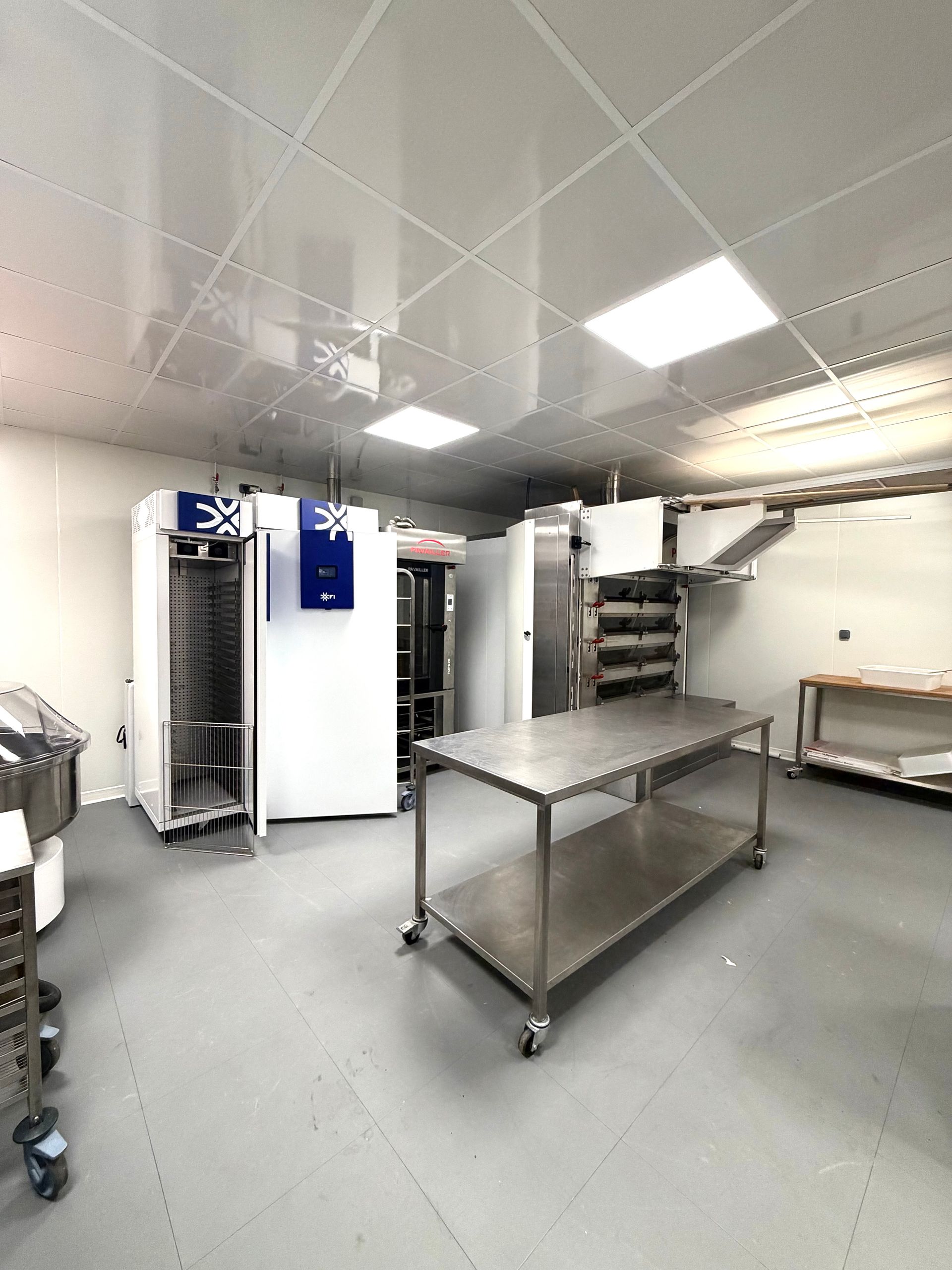 Une cuisine commerciale stérile aux murs blancs, équipée de tables de travail en acier inoxydable, d'unités de refroidissement et d'étagères pour le matériel.