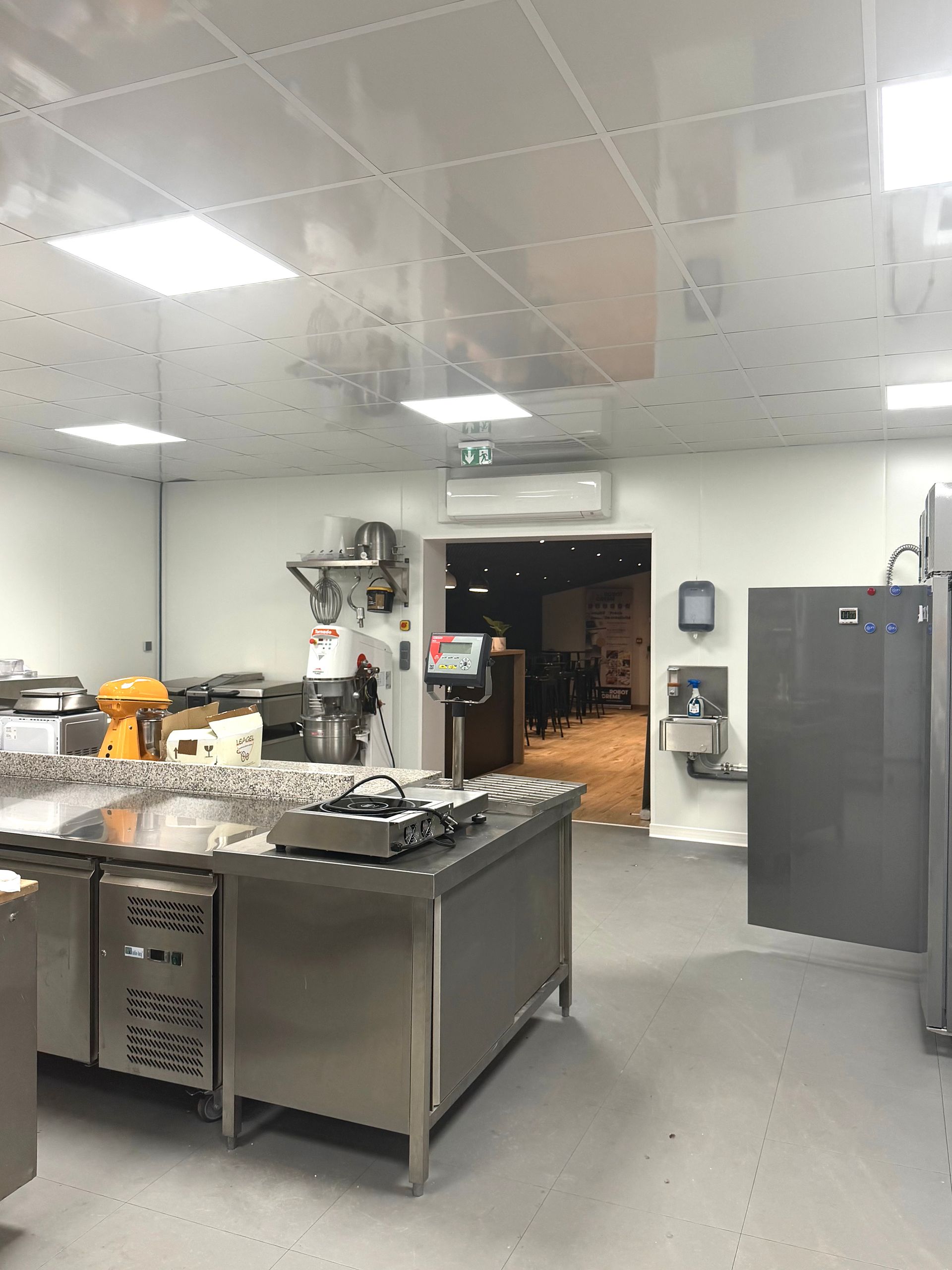 Une cuisine professionnelle propre, avec des postes de travail en acier inoxydable, un grand mélangeur industriel et un plafond carrelé lumineux.