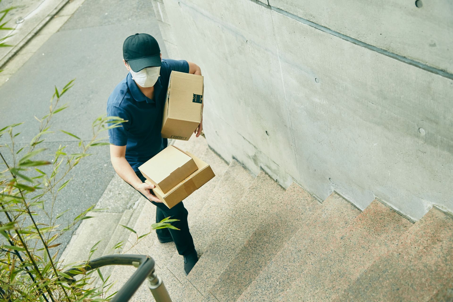 Un livreur portant un masque et une casquette transporte deux cartons en haut d'un escalier.
