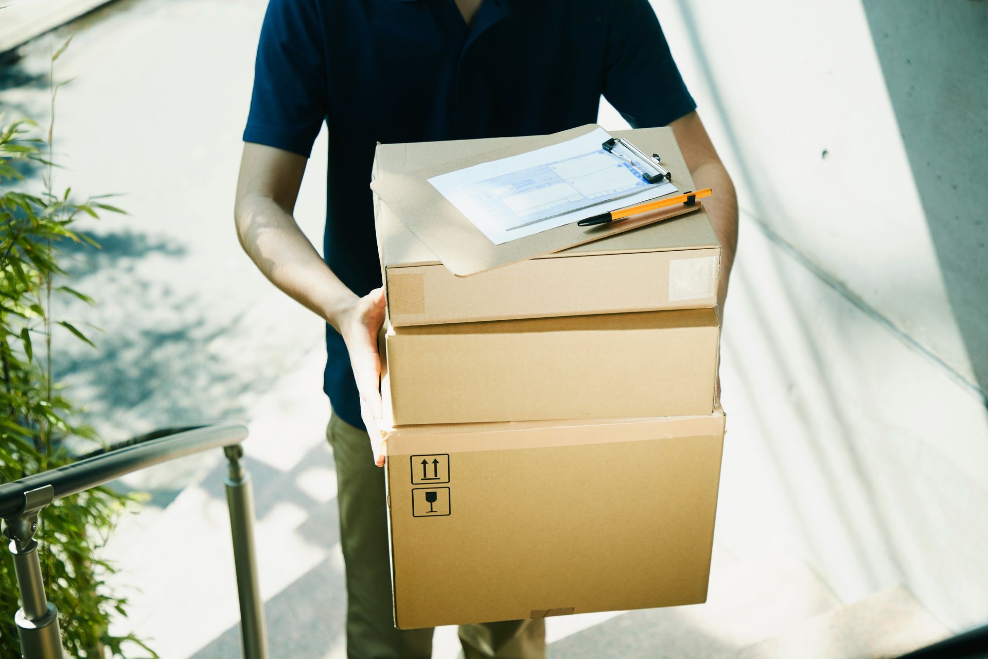 Un livreur en chemise bleu foncé transporte une pile de trois cartons bruns avec un bloc-notes posé dessus.