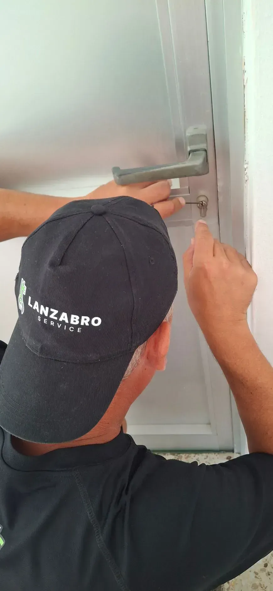 Una persona con gorra y camisa negras instalando o reparando una cerradura en una puerta blanca.