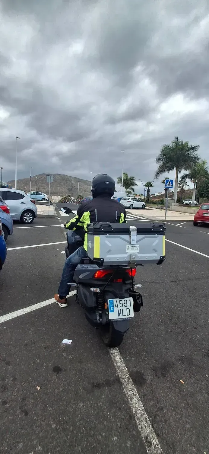 Un motociclista con un chaleco reflectante y una gran caja de carga espera en un estacionamiento bajo un cielo nublado.