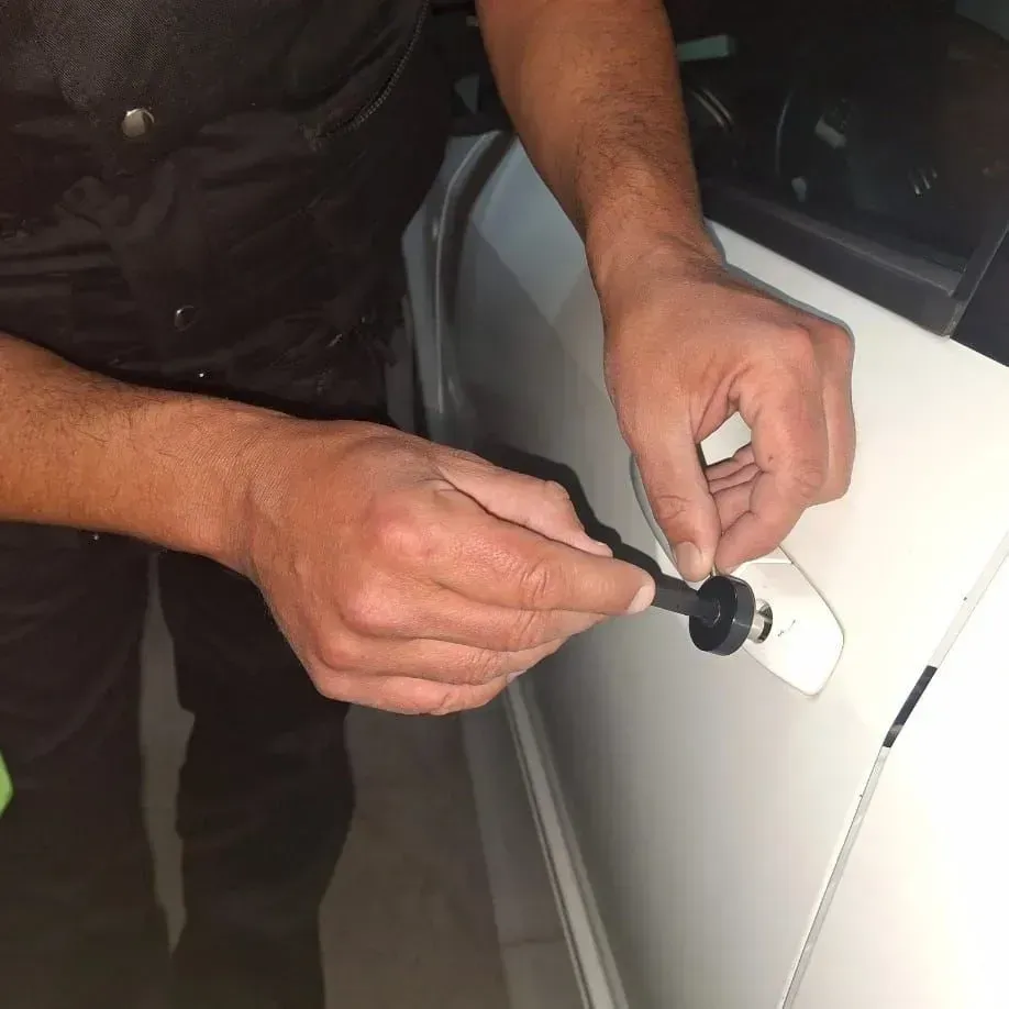 Una persona vestida de oscuro utiliza una pequeña herramienta negra para desbloquear o quitar la manija de la puerta de un coche blanco.