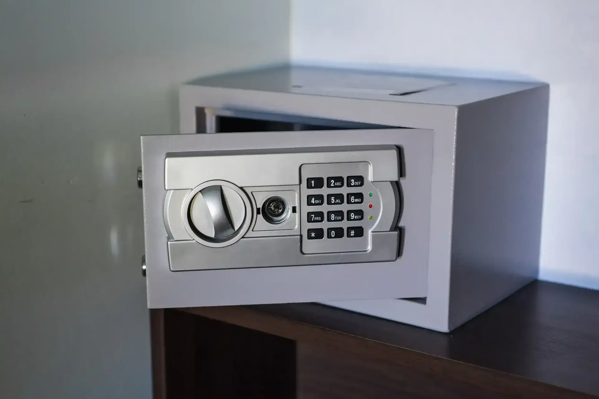 Una caja fuerte digital de color gris, con la puerta abierta, colocada sobre una superficie de madera oscura contra una pared lisa.