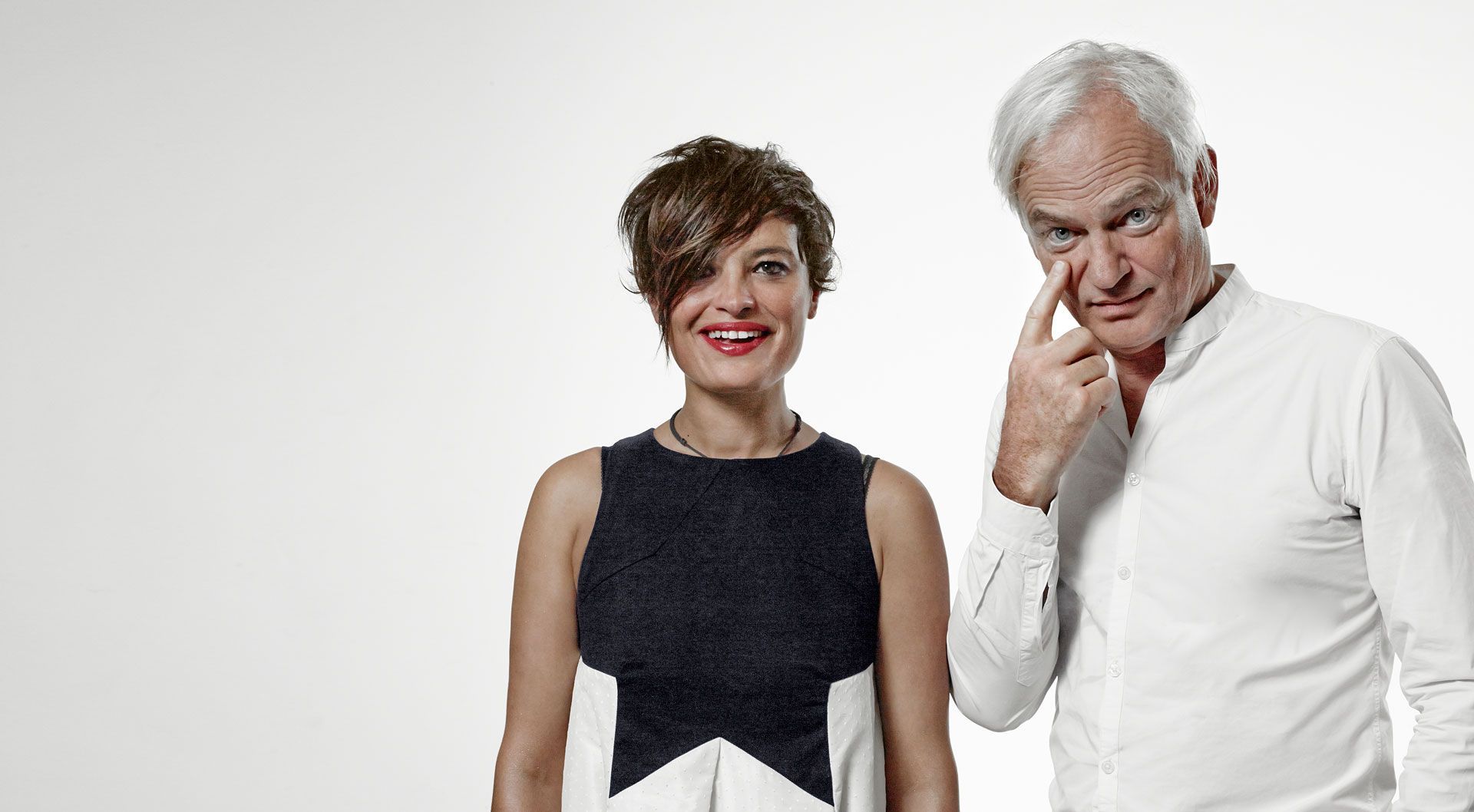 Une femme en robe noire et blanche et un homme avec un doigt sur l'œil, tous deux souriants, sur fond blanc.