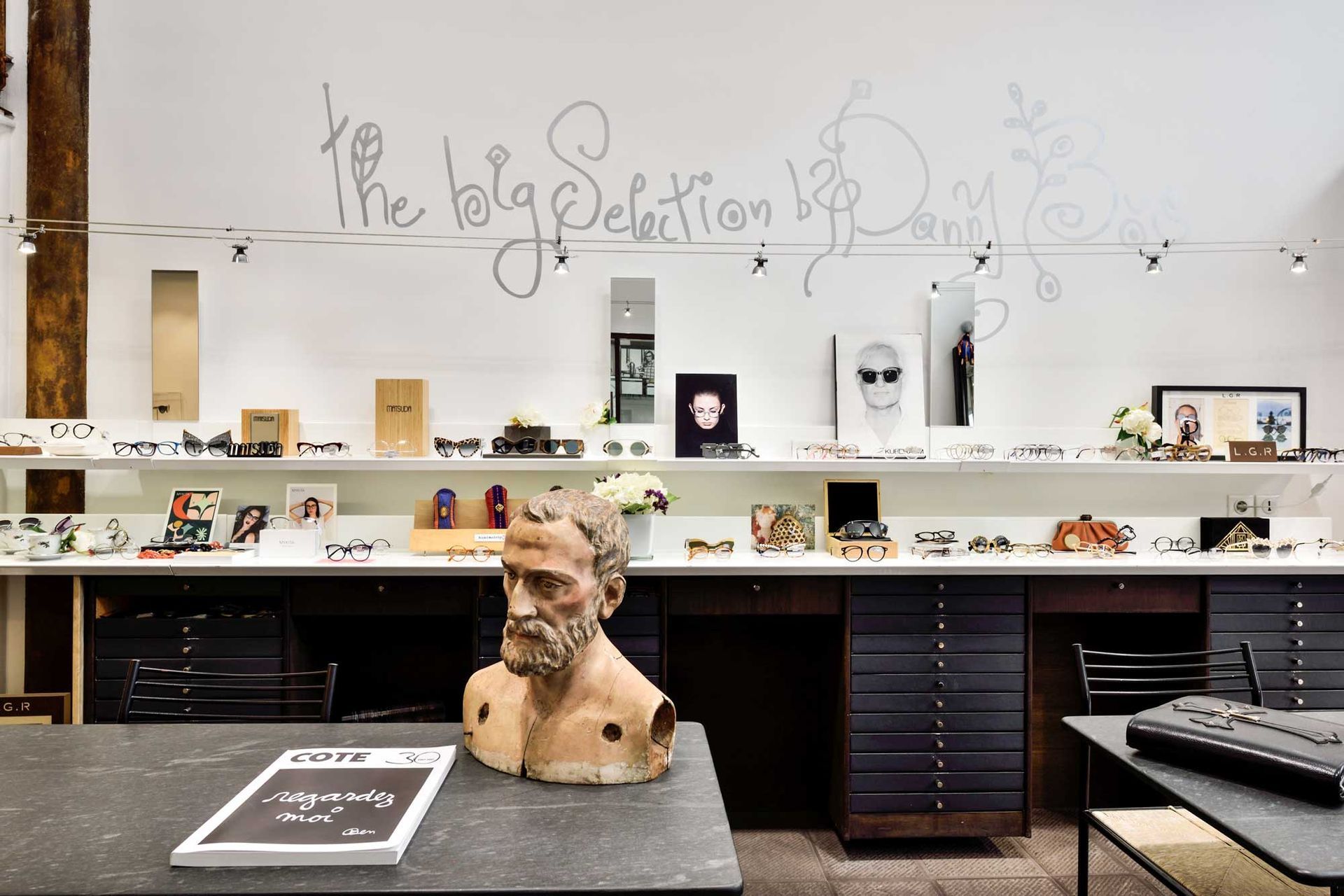 Intérieur d'un magasin de lunettes avec des lunettes exposées sur un comptoir sous un mur blanc avec un texte manuscrit et un buste.