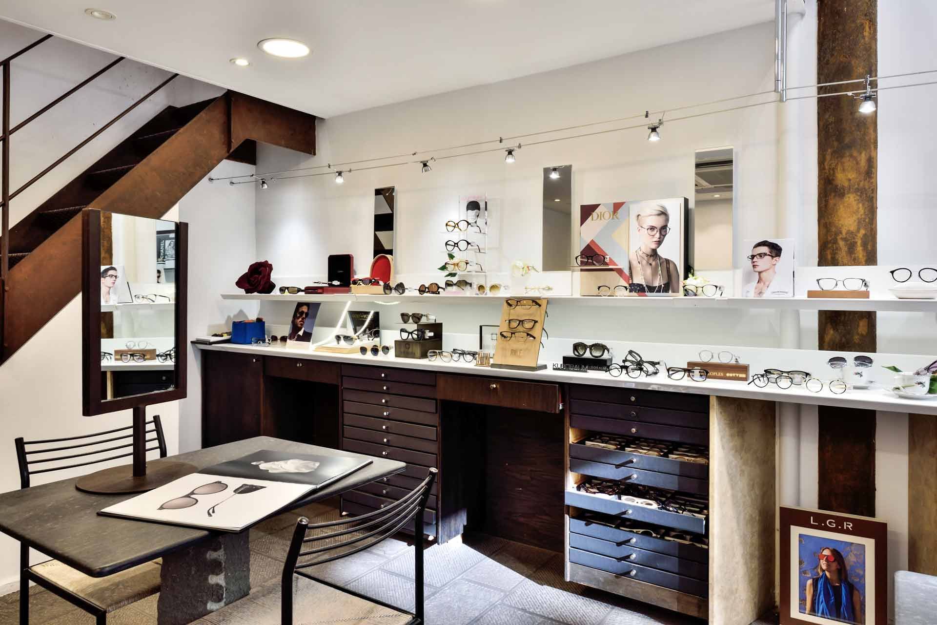 Intérieur d'un magasin de lunettes : exposition de montures, table avec dessins, escalier et miroirs.
