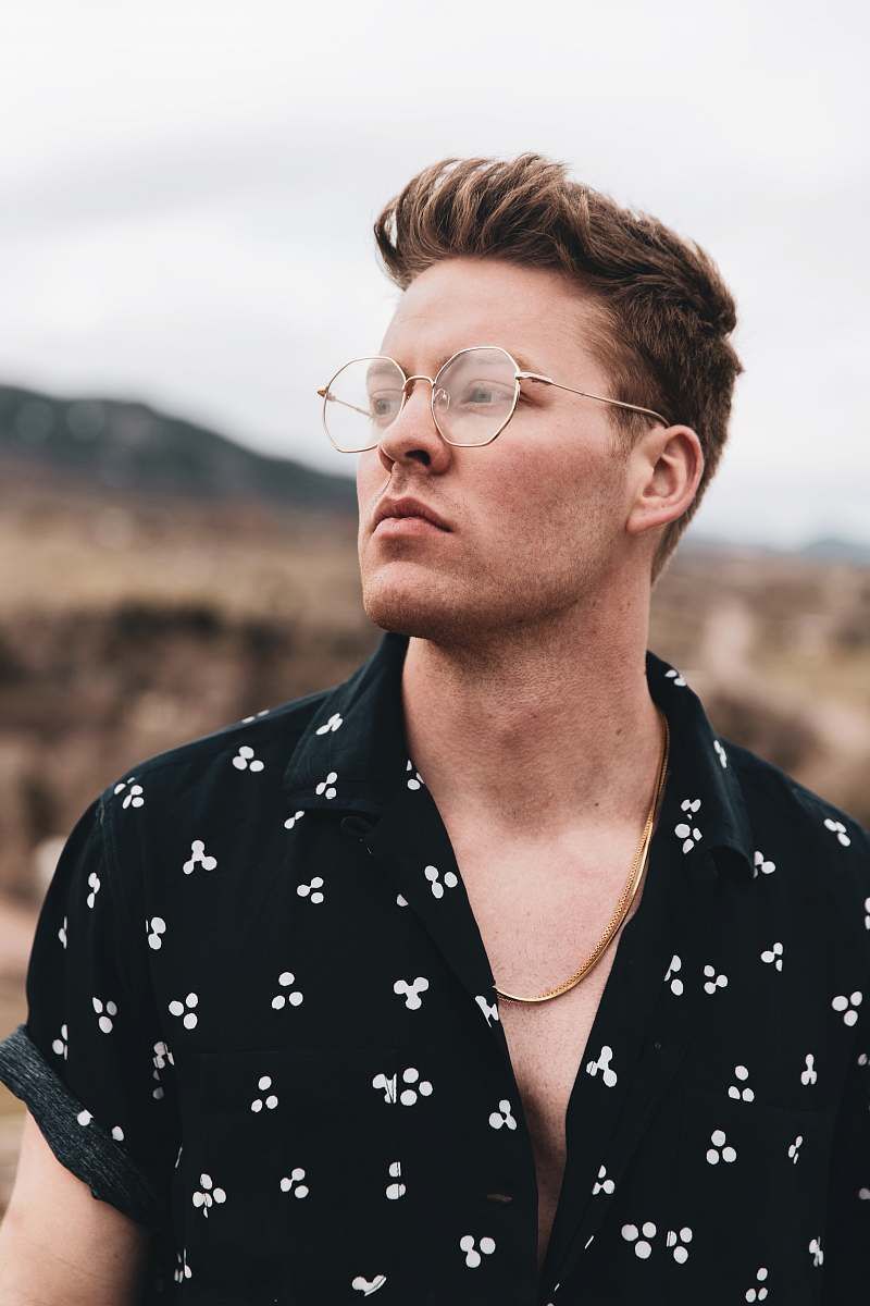 Un homme portant des lunettes et une chaîne en or détourne le regard, vêtu d'une chemise à motifs dans un décor extérieur.