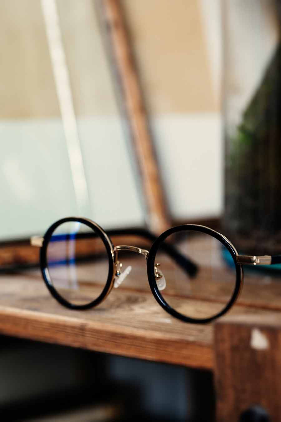 Lunettes noires à monture ronde sur une étagère en bois. Touches dorées et verres transparents.