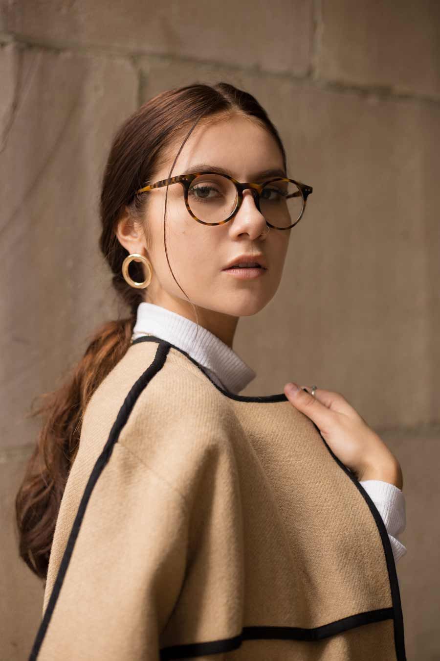 Femme portant des lunettes et des créoles dans une cape beige et noire, regardant par-dessus son épaule.