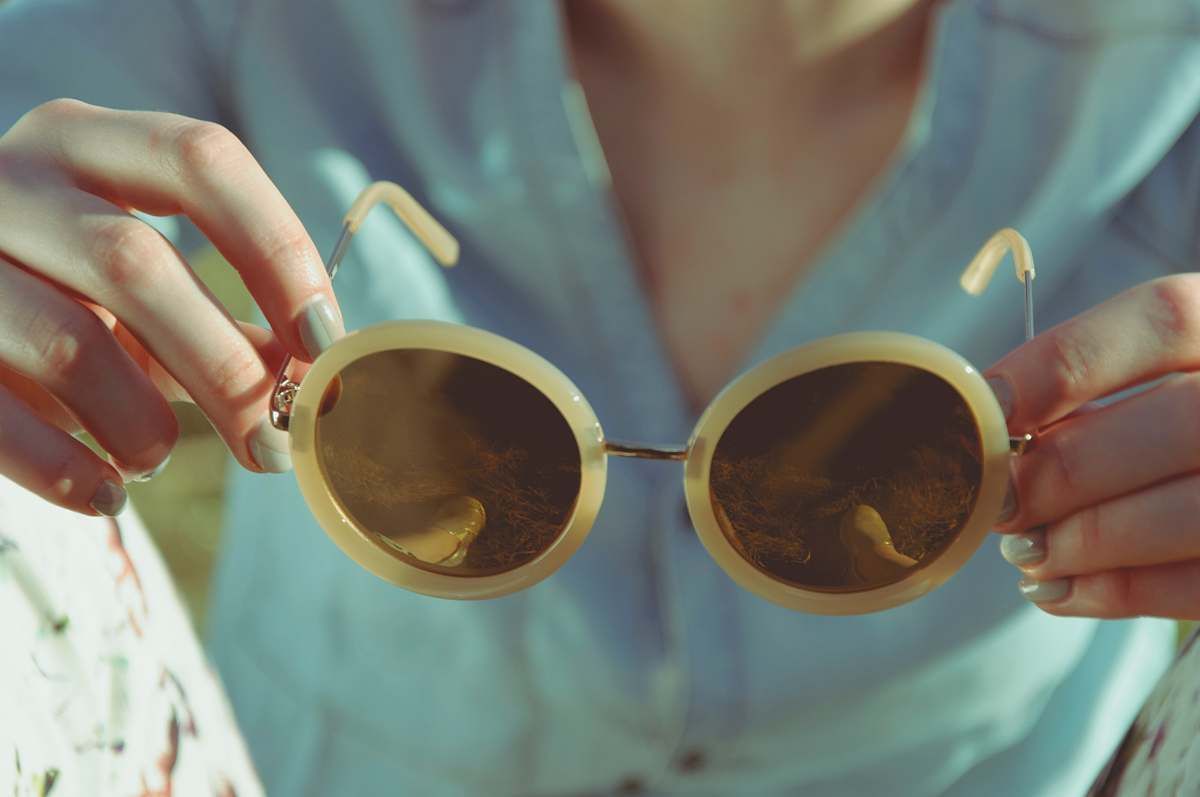 Mains tenant des lunettes de soleil rondes de couleur crème avec des verres marron.
