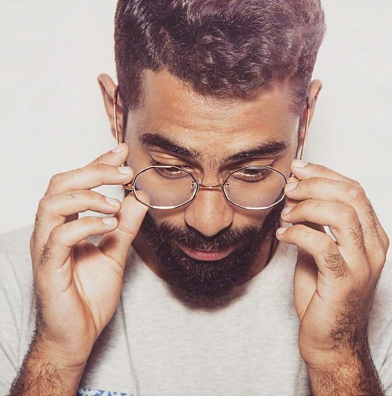 Homme avec une barbe et des lunettes les ajustant avec les deux mains ; fond clair.