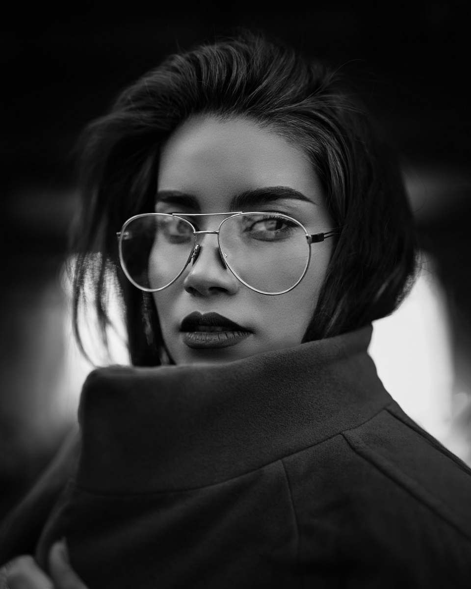 Femme portant des lunettes et un manteau, regardant vers l'objectif, l'air sérieux. Photo en noir et blanc.