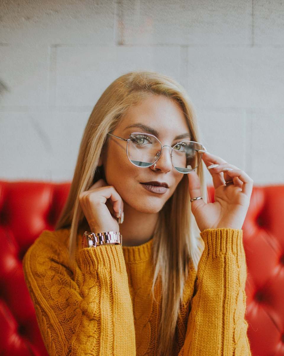 Une femme blonde en pull jaune ajuste ses lunettes, assise sur un canapé rouge.