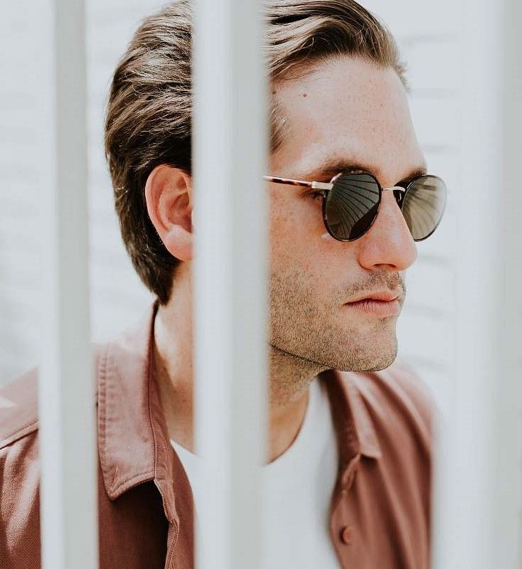 Homme portant des lunettes de soleil, tourné vers la droite, encadré de barres blanches. Il porte une chemise rose et un t-shirt blanc.