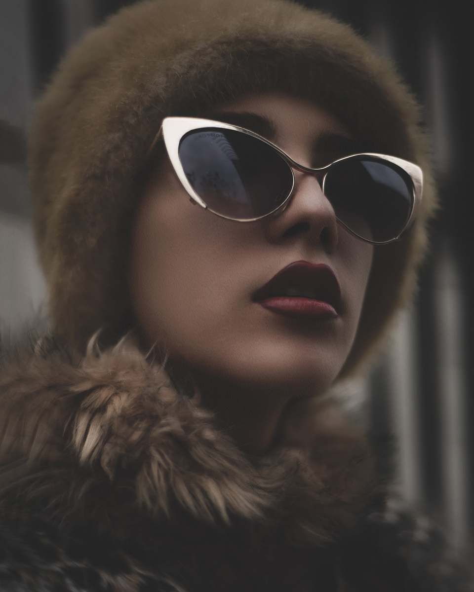 Femme portant un chapeau de fourrure et des lunettes de soleil, levant les yeux, rouge à lèvres foncé.