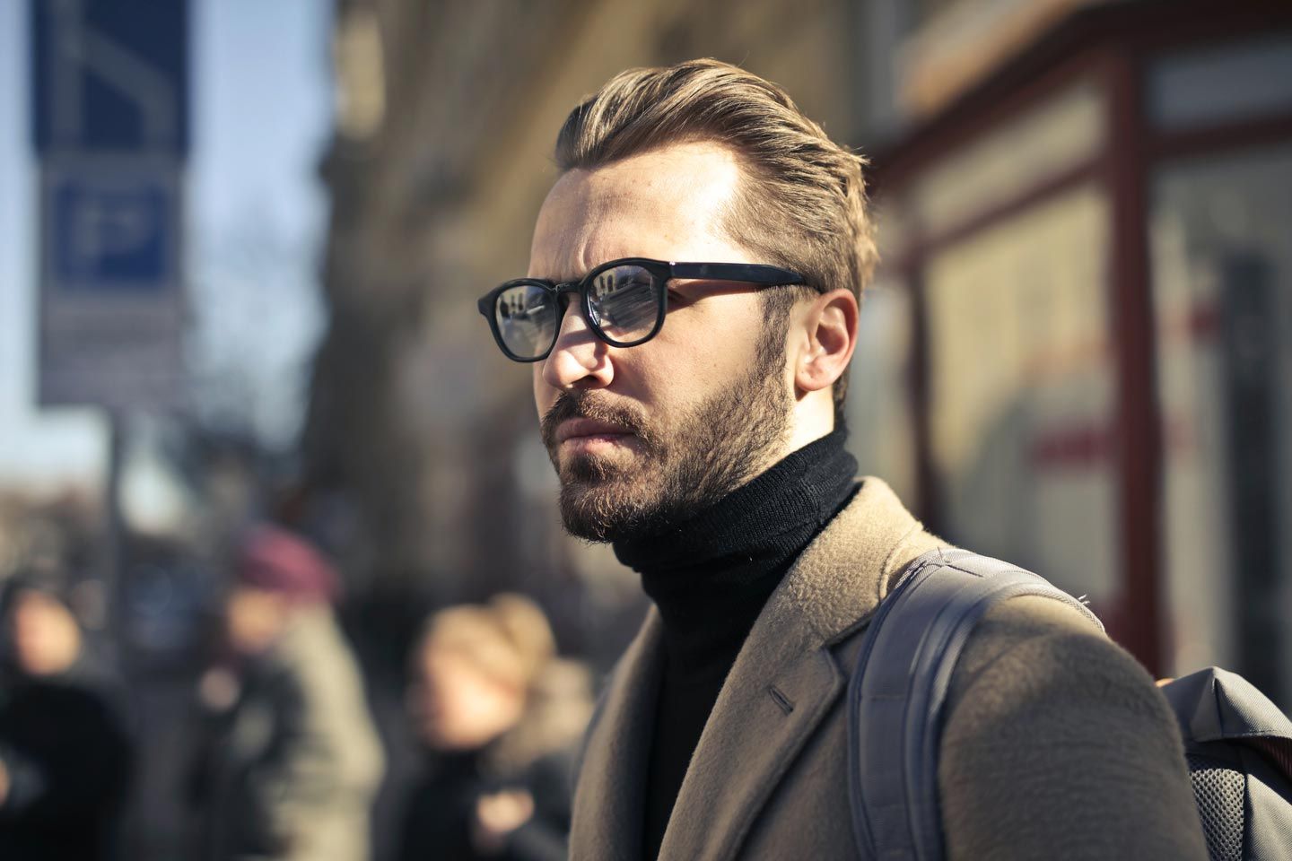 Homme avec des lunettes et une barbe, portant un col roulé et un manteau noirs, regardant vers le côté à l'extérieur.