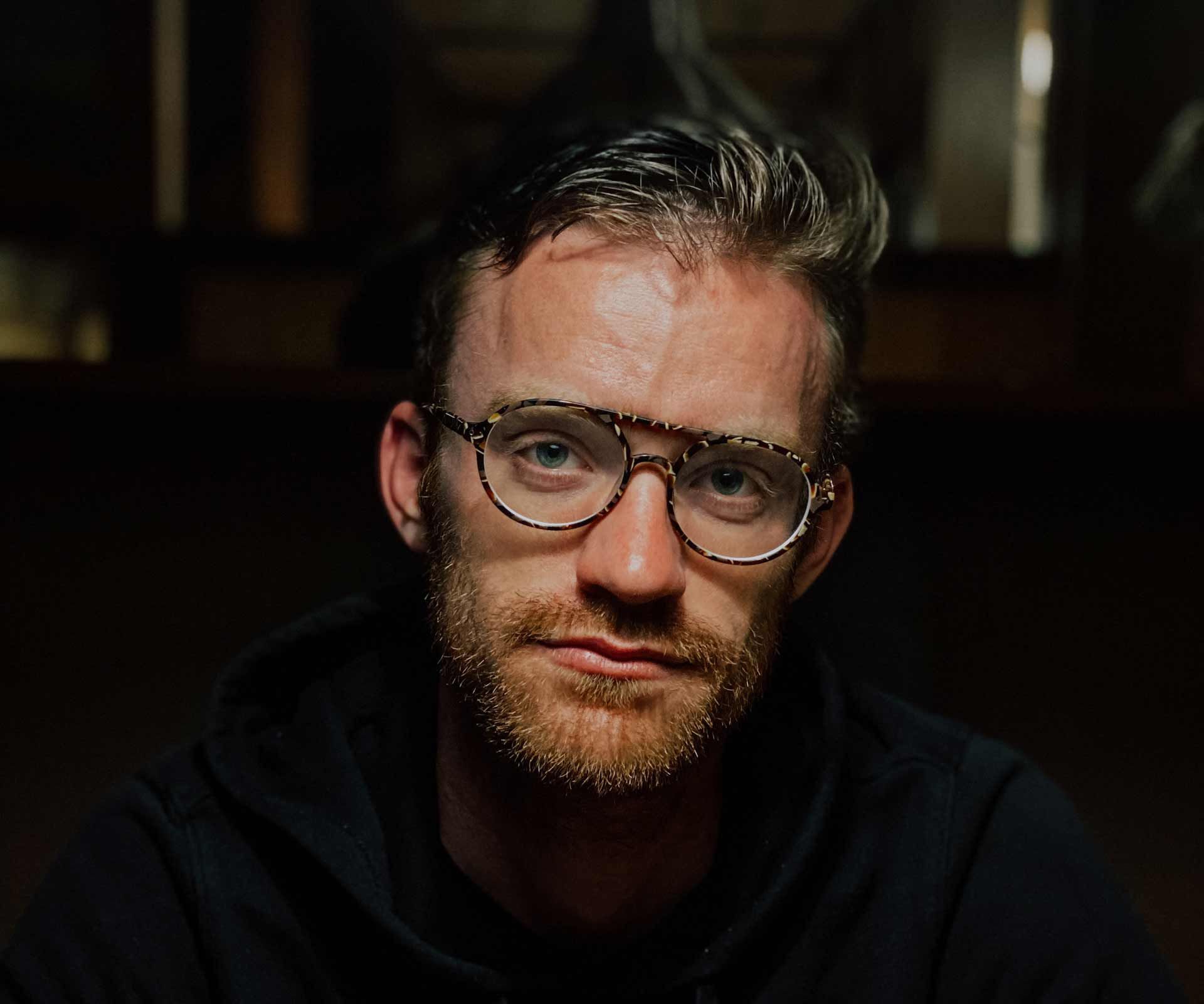 Homme à la barbe rousse et aux lunettes, regardant droit devant lui. Fond sombre.