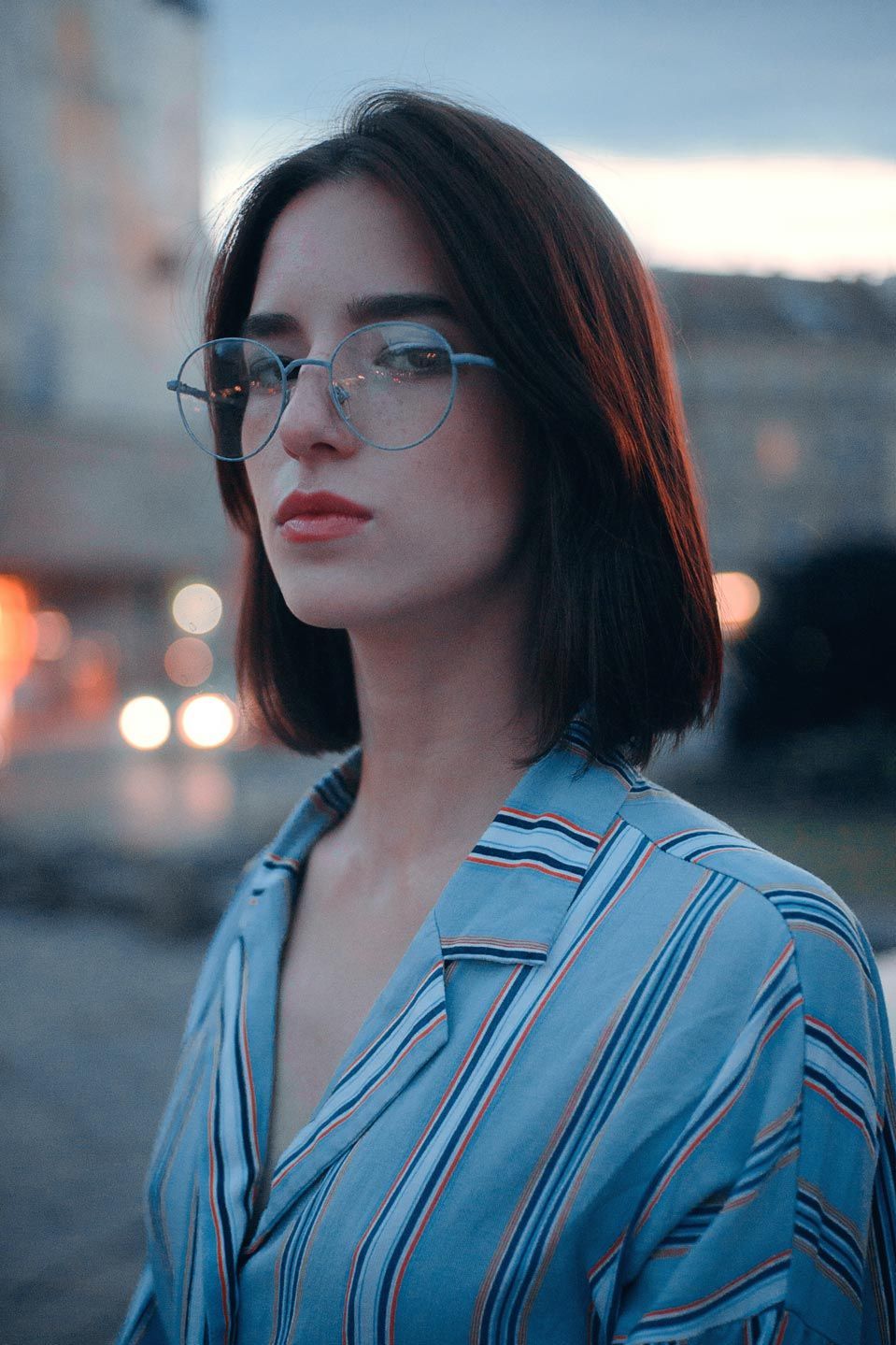 Femme avec une coupe au carré et des lunettes portant une chemise bleue rayée, à l'extérieur avec les lumières floues de la ville.