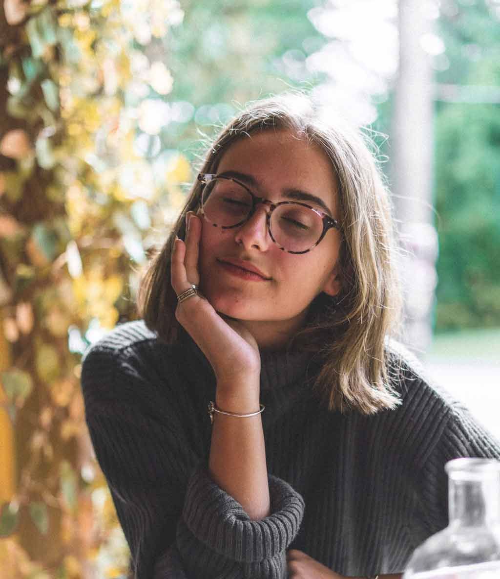 Femme portant des lunettes, la tête appuyée sur sa main, les yeux fermés, l'air détendu. Extérieur, feuillage en arrière-plan.