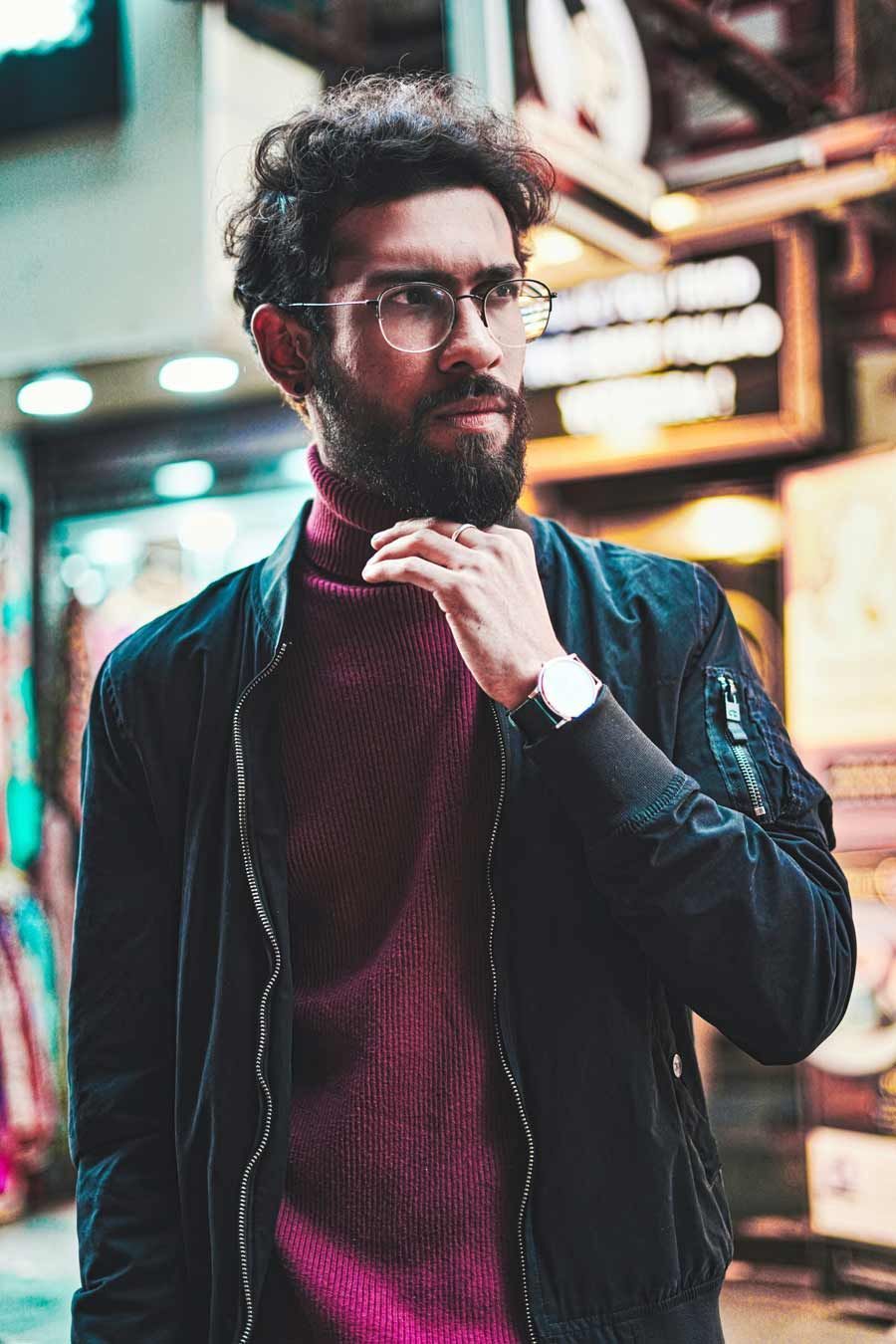 Homme avec des lunettes et une barbe, portant une veste bomber noire sur un col roulé bordeaux, dehors la nuit.