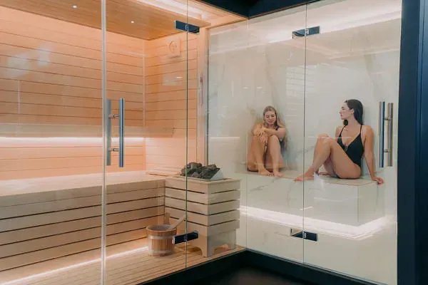 Dos mujeres están sentadas en una sauna con puertas de cristal.