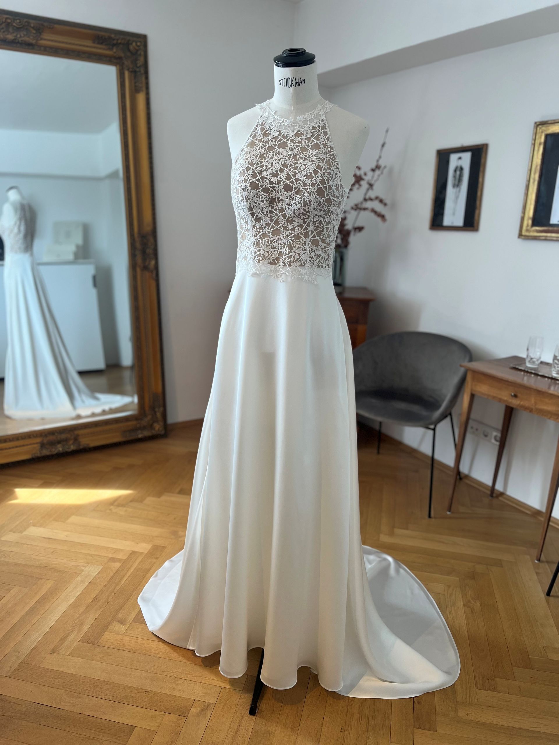 Weißes Brautkleid an einer Schaufensterpuppe in einem hellen Raum, mit Oberteil aus Spitze und fließender Schleppe.
