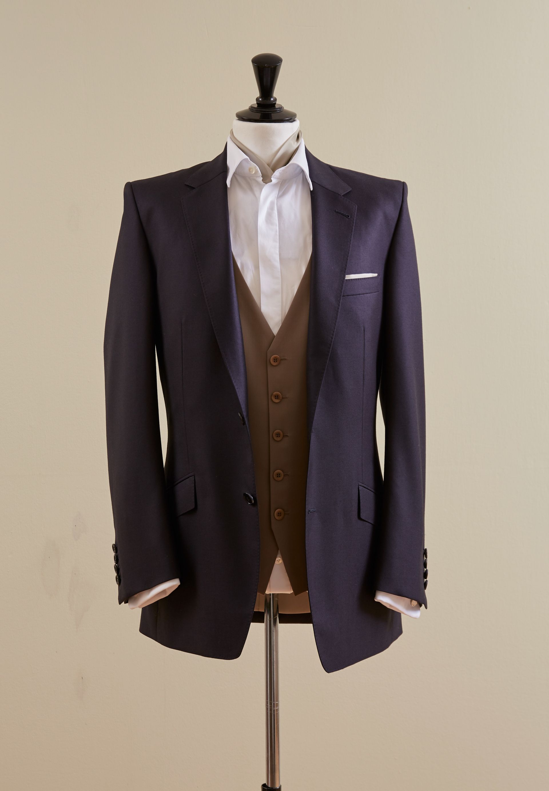 Schaufensterpuppe mit marineblauem Blazer, brauner Weste und weißem Hemd vor beigem Hintergrund