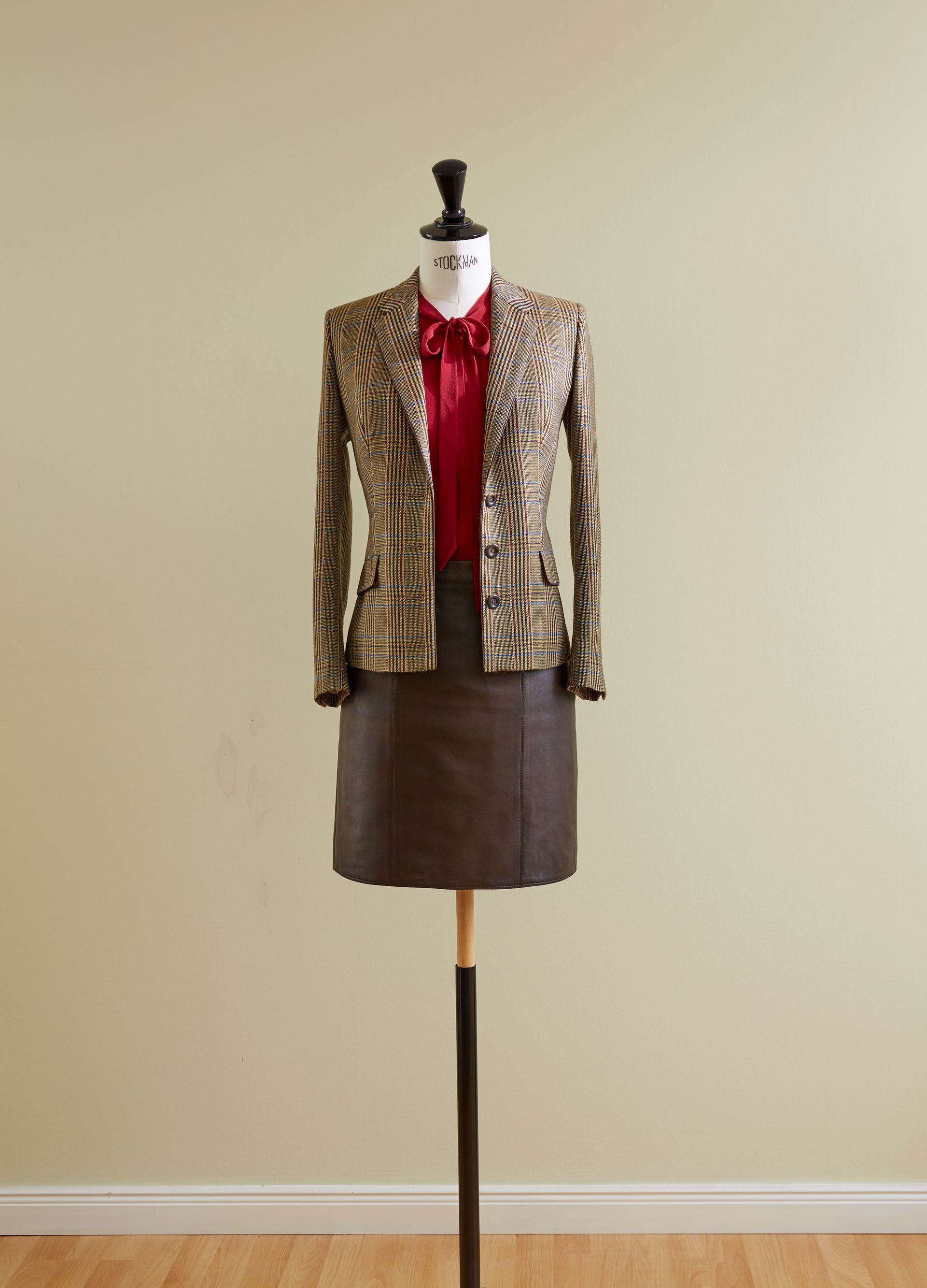 Schaufensterpuppe mit gemusterter Jacke, roter Bluse und braunem Rock vor beiger Wand.