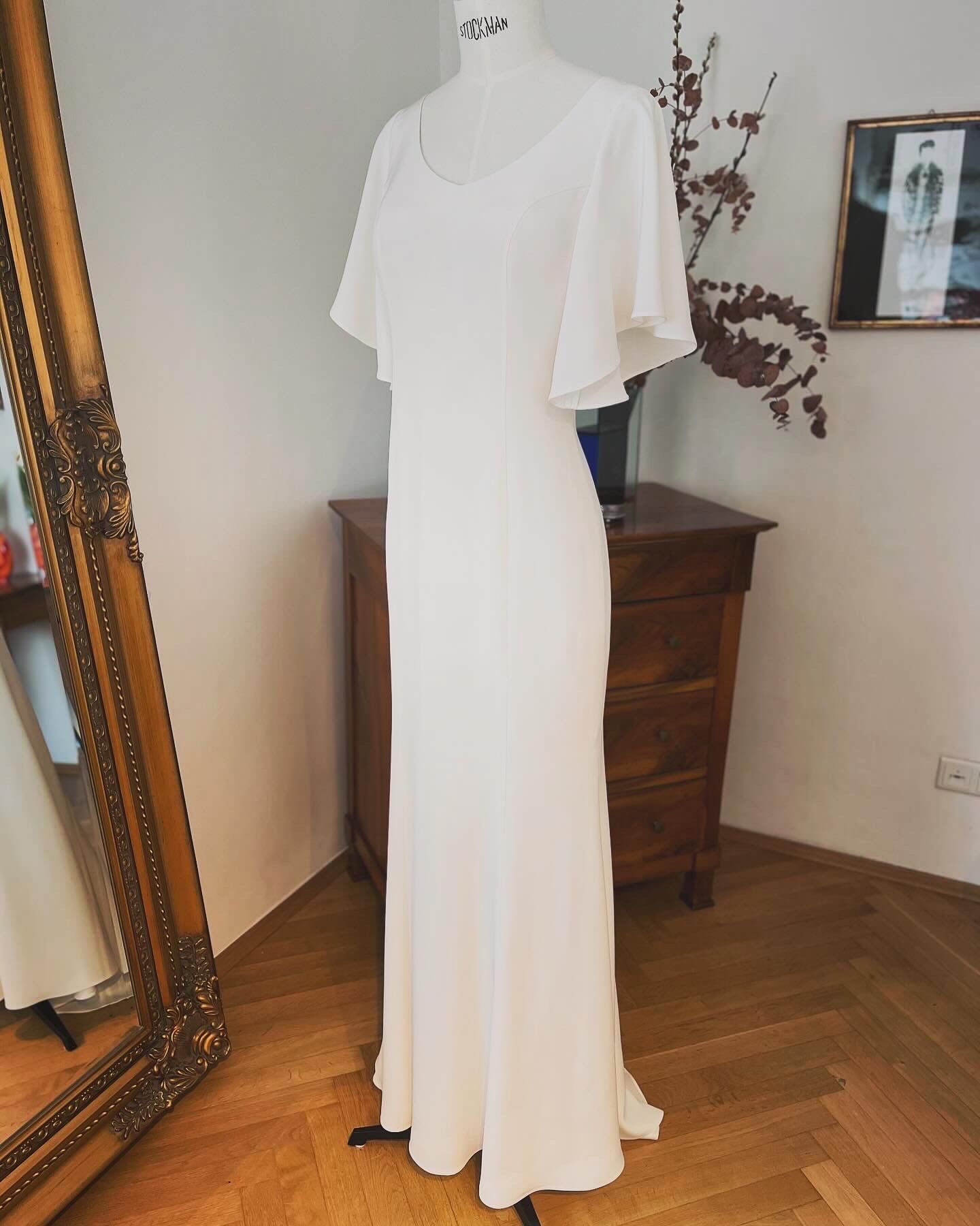 Ein weißes, bodenlanges Kleid hängt auf einer Schaufensterpuppe in einem Zimmer mit einer Holzkommode und einem Spiegel.