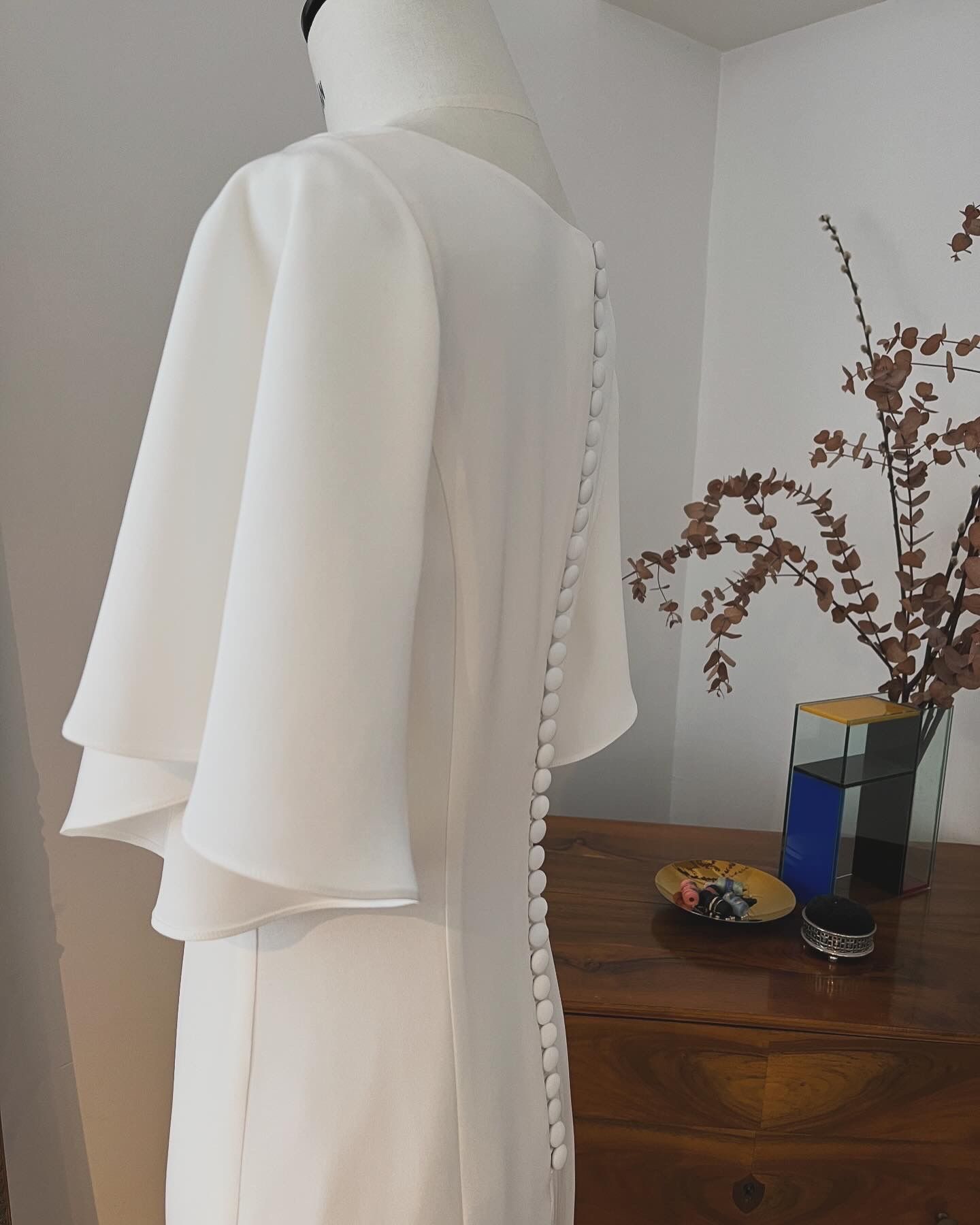 Ein weißes, capeartiges Kleidungsstück hängt auf einer Schaufensterpuppe neben einer Kommode mit einer Vase mit Blumen.