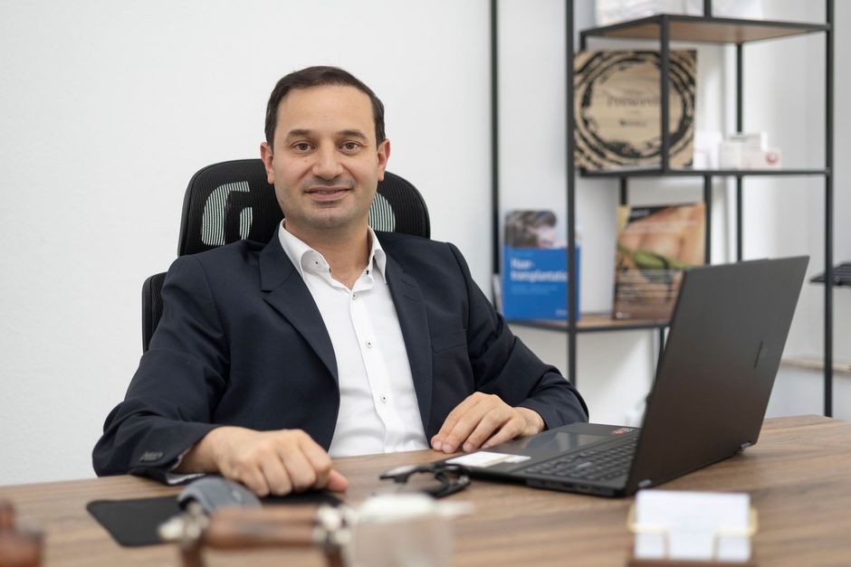 Ein Mann in einem dunkelblauen Blazer sitzt mit einem Laptop an einem Schreibtisch in einem modernen Büro