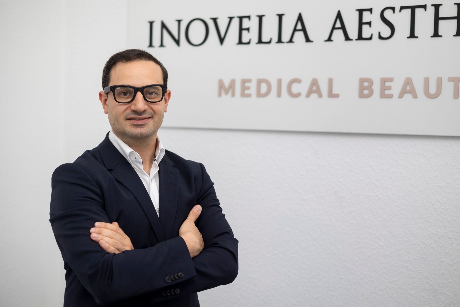 Ein Mann mit Brille und dunkelblauem Anzug steht mit verschränkten Armen vor einer ästhetischen Schönheitsklinik.