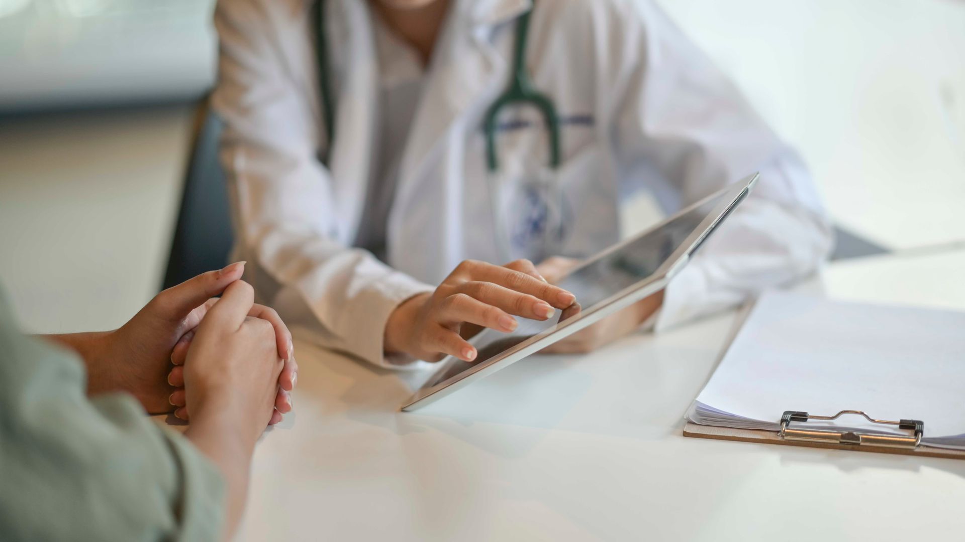 Die Hand eines Patienten wird von einem Arzt neben einem Tablet und einem Klemmbrett in einem Behandlungszimmer gehalten.