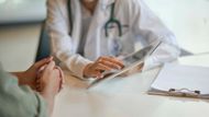 In einer hellen Klinik berät sich ein Arzt mit einem Patienten, der ihm gegenüber am Schreibtisch sitzt und ein Tablet hält.