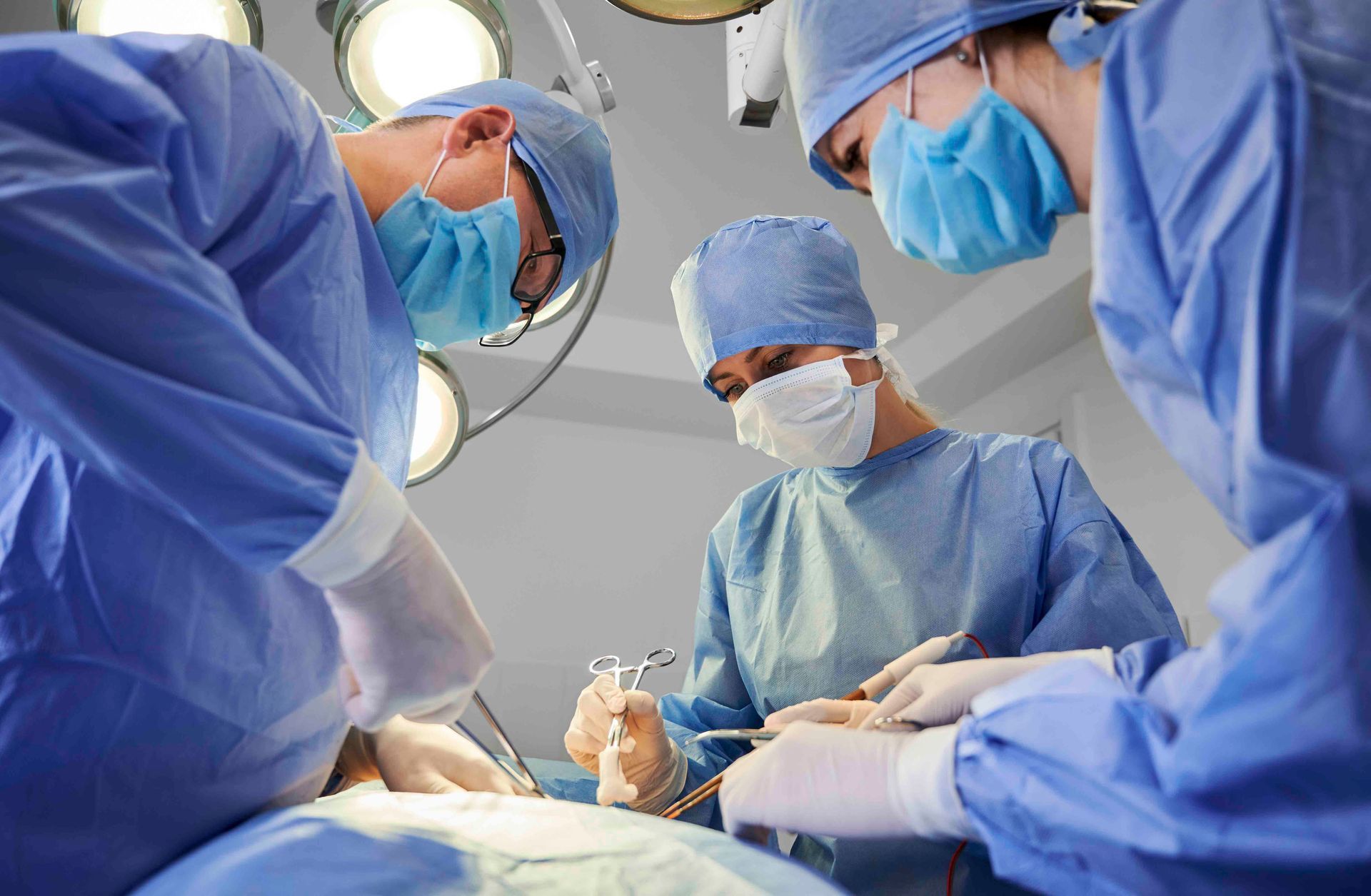 Chirurgen in blauen OP-Kitteln und Masken operieren in einem Operationssaal unter hellem Licht