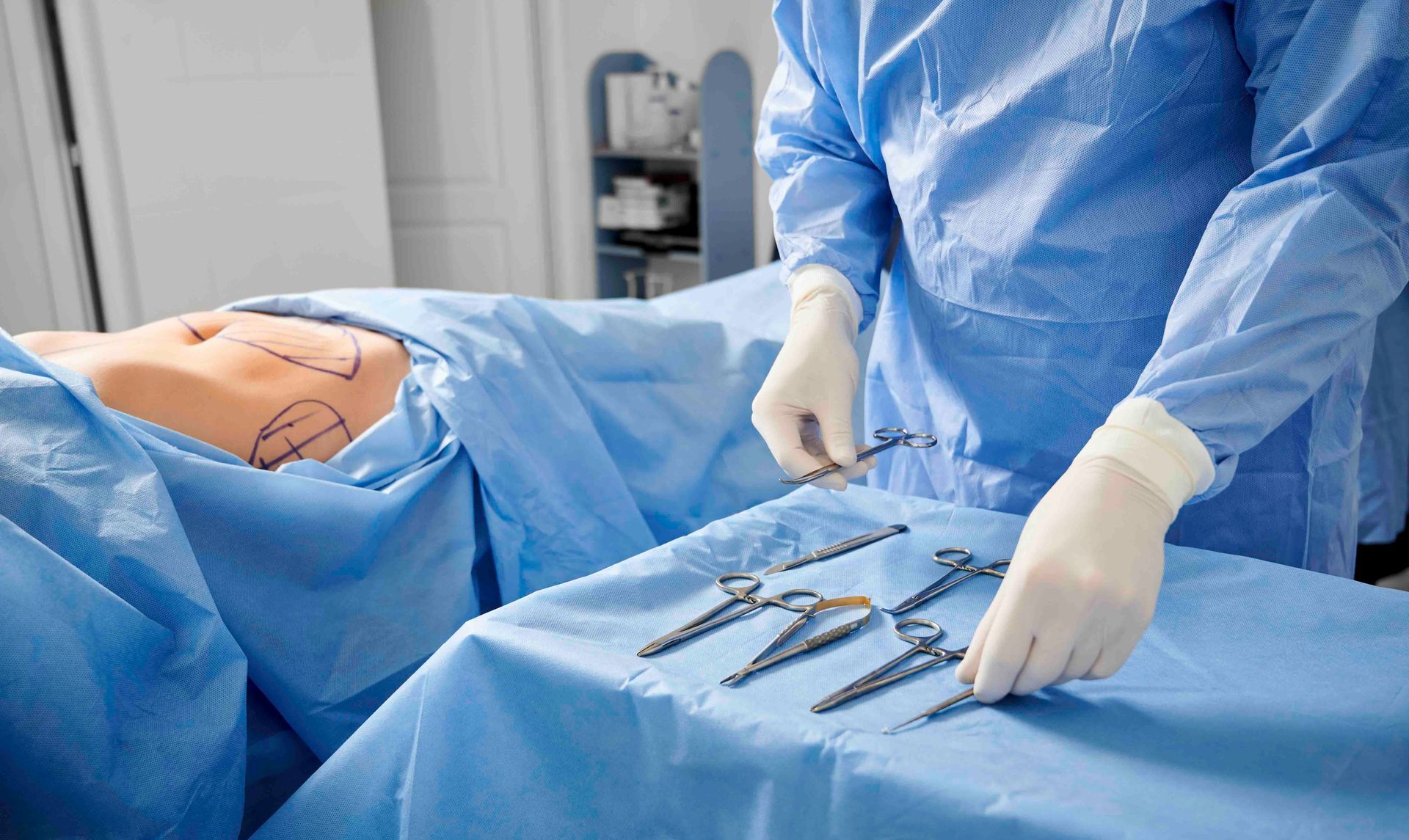 Chirurg in blauer OP-Kleidung arrangiert chirurgische Instrumente neben einem abgedeckten Patienten in einem Operationssaal.