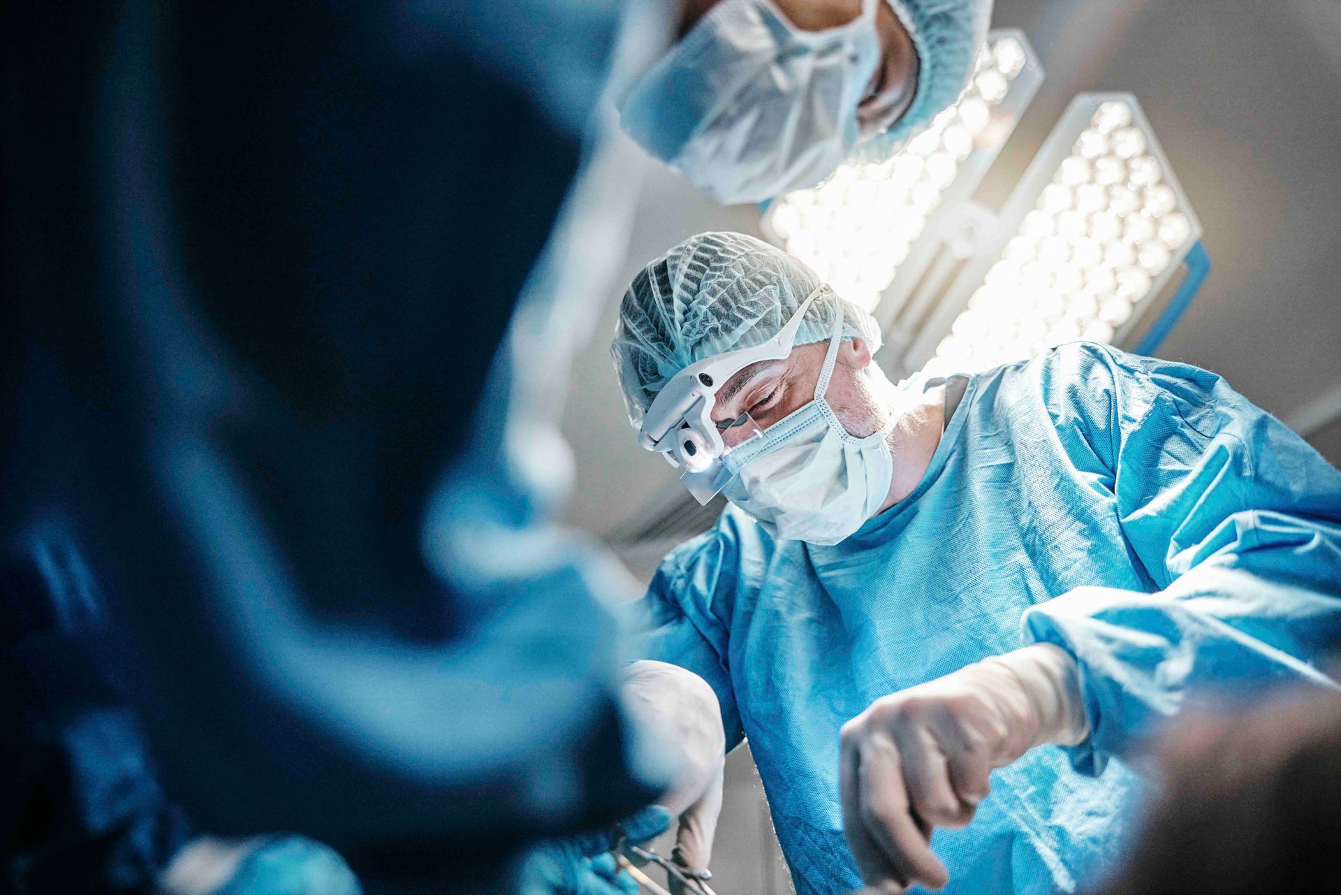 Chirurgen in blauen OP-Kitteln und Masken führen eine Operation unter hellem OP-Licht durch.