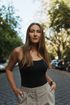 Eine Frau steht im Freien auf einer von Bäumen gesäumten Straße. Sie trägt ein schwarzes Oberteil und beige Hosen.
