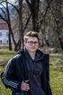 Person mit Brille und Rucksack steht im Freien in einem Park; im Hintergrund sind Bäume und ein Haus zu sehen.
