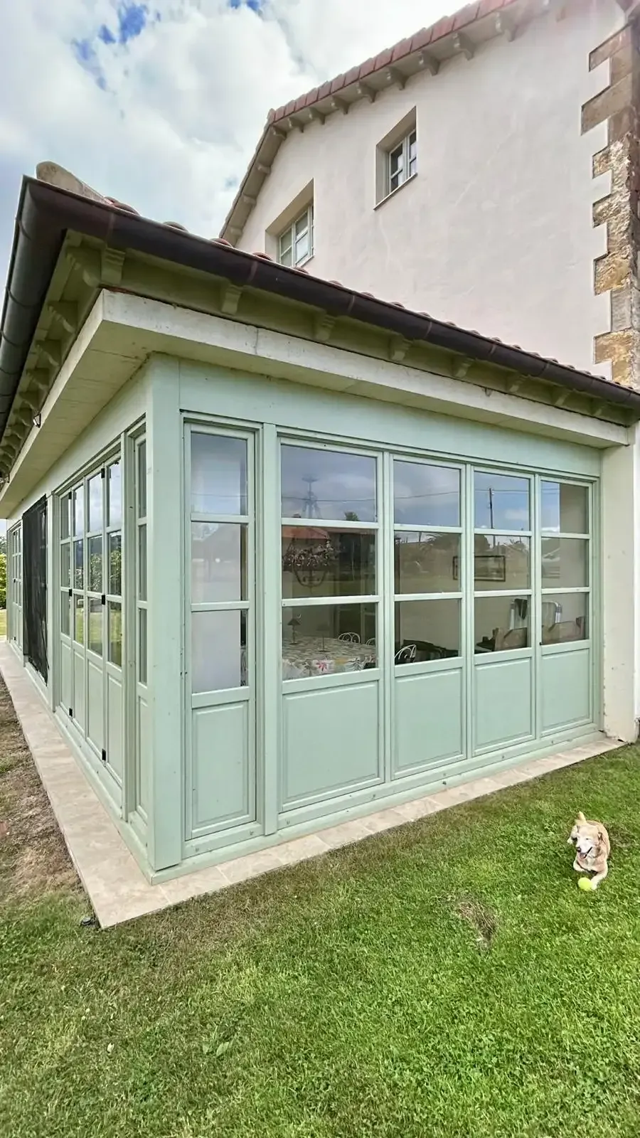 Ampliación de una terraza acristalada de color verde claro en un edificio de color claro con grandes ventanales y césped.