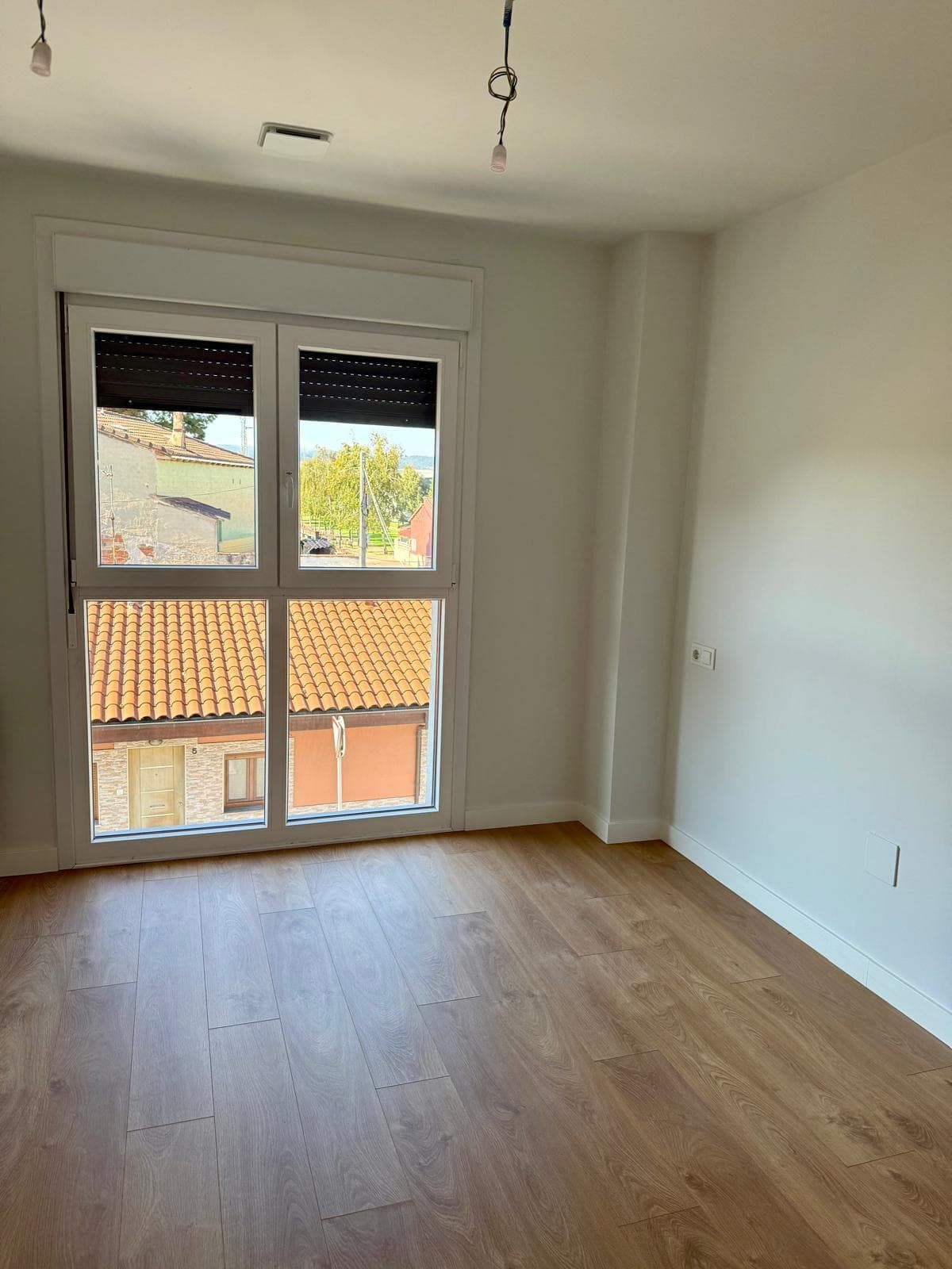 Habitación vacía con suelo de madera y gran ventanal con vistas a los tejados.