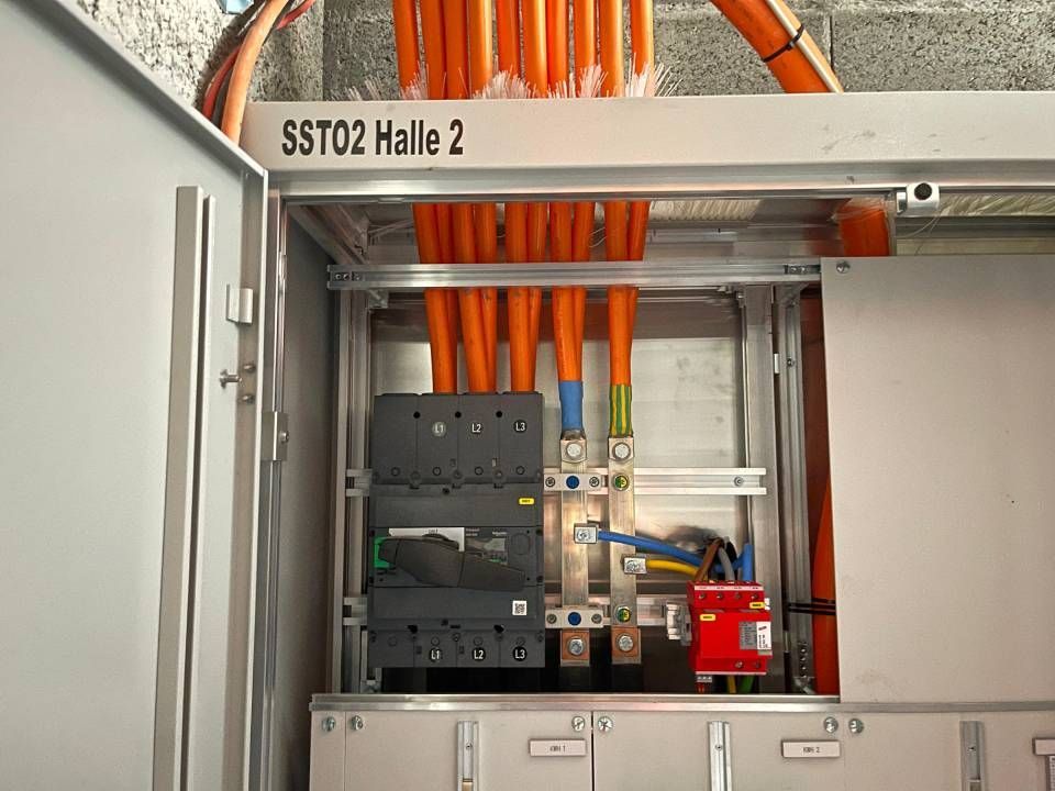 Armoire électrique avec câbles orange, disjoncteurs et barres omnibus. Étiquette « SST02 Halle 2 » visible.