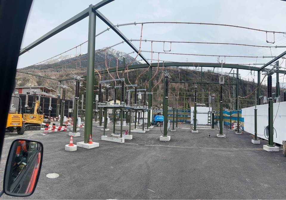 Un poste de transformation électrique en construction, avec des structures métalliques, des câbles et une petite excavatrice. Montagnes en arrière-plan.