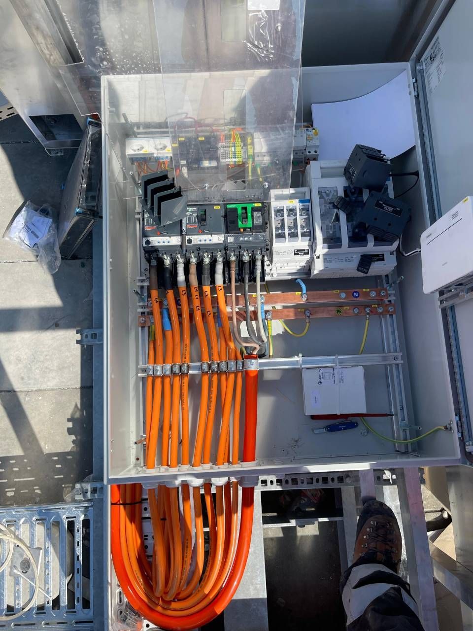 Tableau électrique avec câbles orange, composants divers et couvercle transparent, installé à l'extérieur.